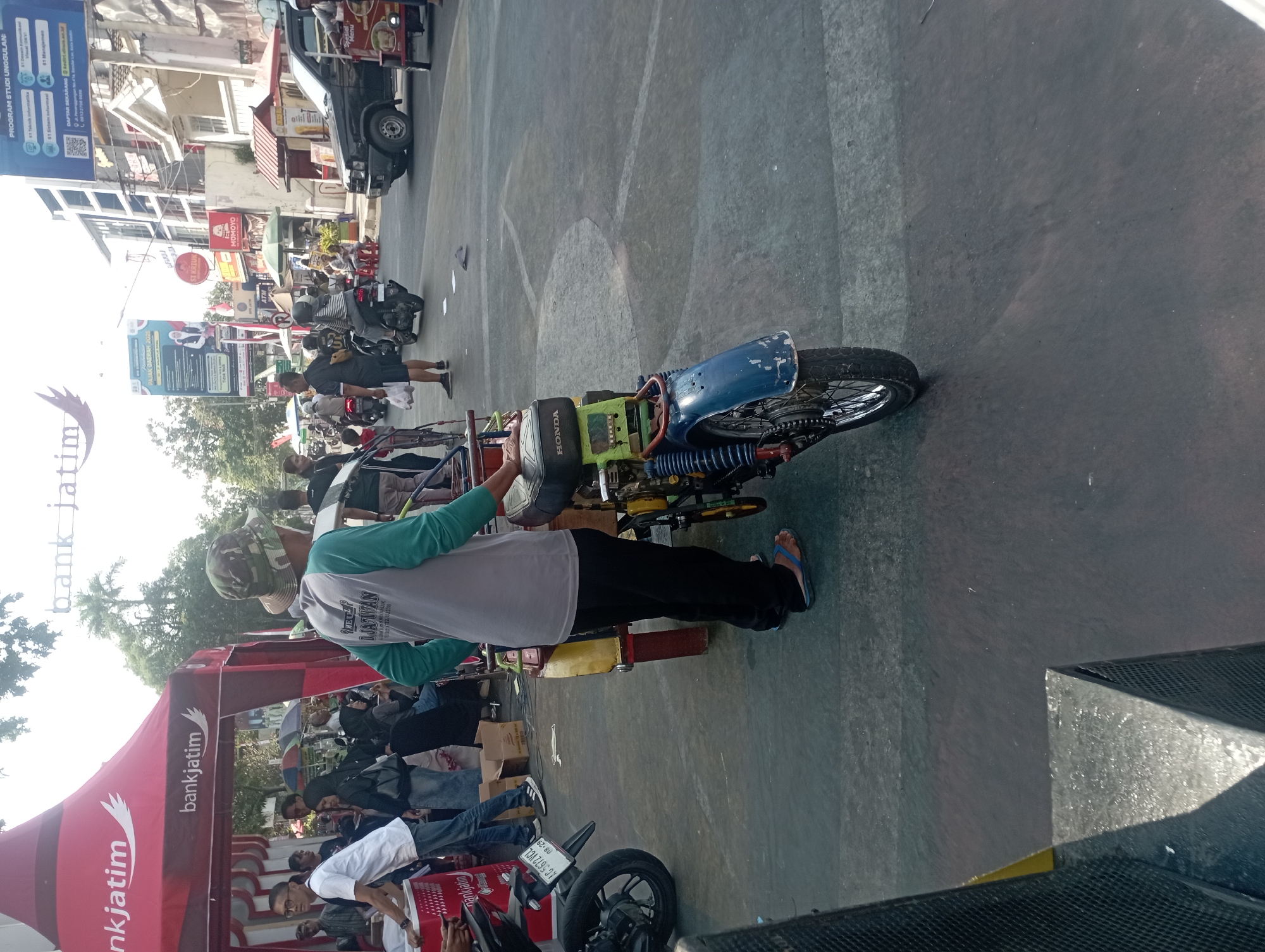 The image depicts a street scene in Nganjuk, Indonesia, on a sunny day. In the foreground, a person wearing a camouflage hat, a grey and teal long-sleeved shirt, and black pants stands next to a modified motorcycle with a sidecar. The motorcycle has a blue fender, a visible chain, and the "HONDA" brand name on its fuel tank. The sidecar appears to be made of yellow metal. The person is wearing flip-flops.
Behind the person and the motorcycle, a red tent with "bankjatim" branding is set up, suggesting a promotional event. Several other people are visible, some standing and interacting near the tent and a white table. One individual in a white t-shirt and blue jeans is partially visible to the left. Other people are seen in the background, some on motorcycles.
The street is paved with asphalt, and there are white painted lines on the road. Buildings line the street, including one with multiple stories and prominent advertising signs. One sign displays "PROGRAM STUDI UNGGULAN" and mentions various IT-related programs. Another sign shows "STIKOM" and an image of a bear. A "MOMOYO" restaurant sign is also visible. Cars and motorcycles are present in the middle ground and background, indicating moderate traffic. The overall atmosphere appears to be a busy outdoor event or market.