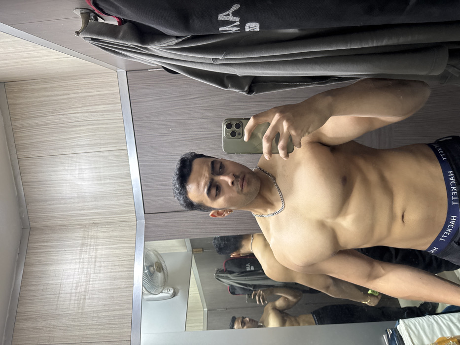 A male individual is depicted in a mirror selfie, shirtless and displaying a muscular physique with prominent pectorals and abdominal definition. He wears dark boxer briefs with "HACKETT" visible on the waistband and a silver chain necklace. His hair is short, dark, and curly, and he has a small earring in his left ear. He holds a light green/gold smartphone, possibly an iPhone, in his right hand, positioned to capture his reflection. His gaze is directed towards the phone/mirror.
The background consists of light-colored, wood-grain patterned wall panels with silver trim, suggesting an indoor setting such as a changing room, gym locker room, or private dressing area. To the right of the subject, grey and dark garments hang from two silver hooks mounted on the wall. The mirror's reflection on the left side of the frame shows the subject's back, a wall-mounted electric fan, and additional hanging garments. Some white fabric and stacked objects are visible in the far left of the reflection. The precise location within Kolkata, India, cannot be determined from the visual evidence.