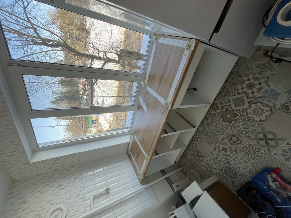 This image depicts an interior room, located in Нарынка, Russia, observed during daytime under clear skies. The room is undergoing renovation or furnishing. The floor is covered with a mosaic of patterned ceramic tiles in shades of brown, grey, blue, and white. The left wall features a lower section of white rectangular tiles, above which is wallpaper with a light background and a subtle leafy pattern. A white radiator pipe with a brass-colored valve extends from the tiled wall.
A large, three-pane window with white frames occupies the central portion of the image, providing a view of an outdoor environment. Outside, patches of snow are visible on the ground, indicating late winter or early spring. Bare deciduous trees and distant evergreen trees are present. A building with a green roof is partially visible in the mid-ground.
Beneath the window, a newly installed wooden-look countertop is visible, spanning the width of the window. Several strips of white masking tape are applied to the countertop surface, and small deposits of a light green substance, consistent with adhesive or sealant, are present at the tape's corners. Below the countertop are white, open base cabinets, some displaying internal shelves. To the far right, a tall, white appliance, possibly a refrigerator, is partially visible. On the floor in the lower-left foreground, a blue bag containing items and additional white cabinet components are present. No individuals are visible within the scene.