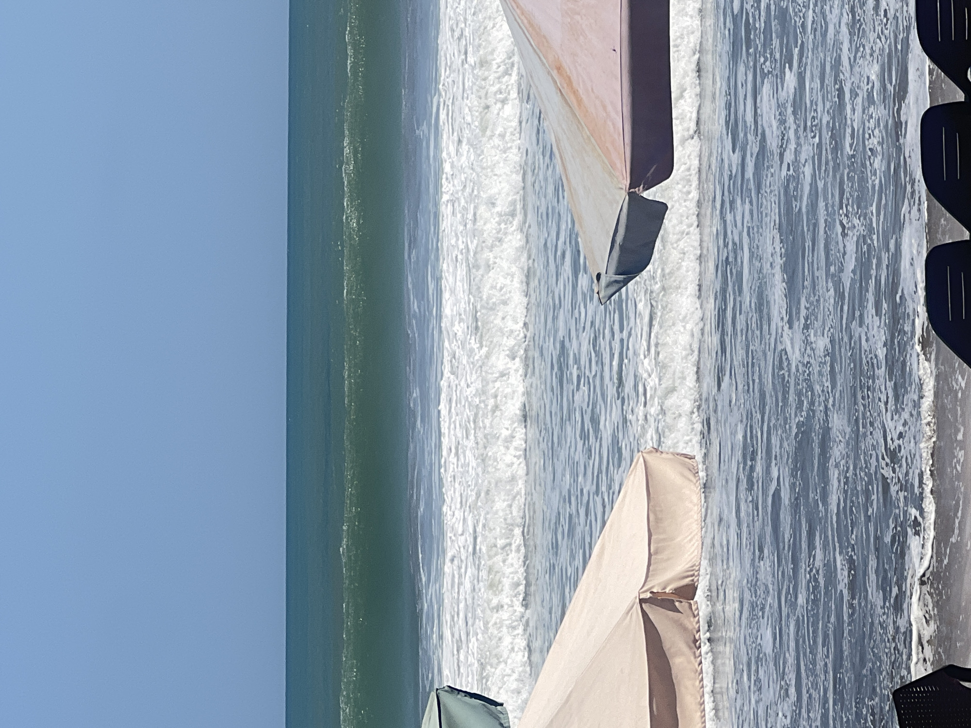 The image shows a view of the ocean at Kuta, Indonesia, with two beach umbrellas in the foreground. The sky is clear and blue. The ocean has waves, and there is white foam on the water. The umbrellas are light-colored, one with a pinkish hue and the other a pale green. Some lounge chairs are partially visible in the bottom of the frame. The scene depicts a typical beach environment in Kuta, known for its surf and popular tourist spots.