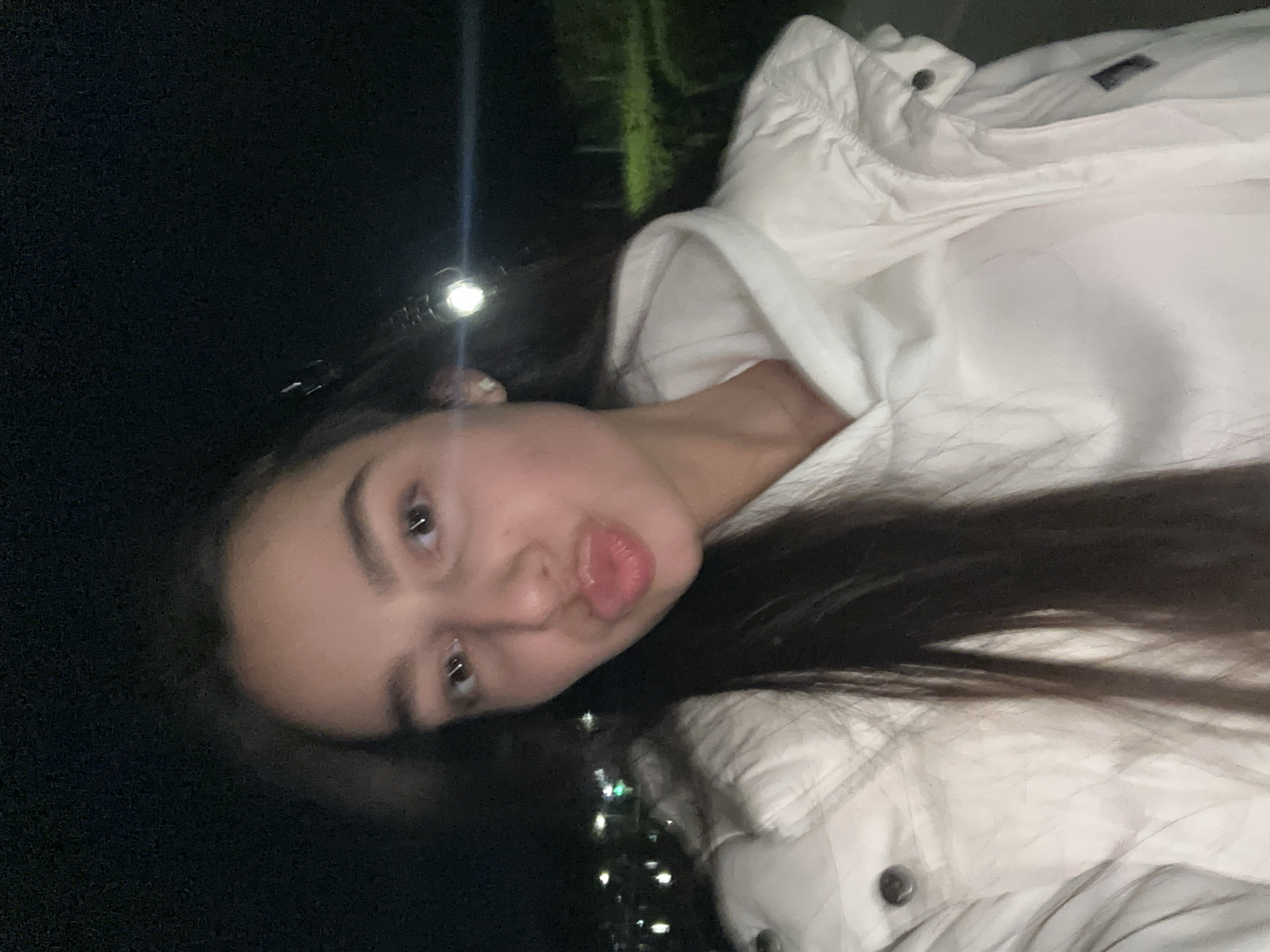 A young woman with long, dark hair and light skin is posing for a selfie outdoors at night in Plovdiv, Bulgaria. She is wearing a white puffy jacket with a hood. Her face is turned slightly to her left and her lips are pursed. The background is dark, with faint streetlights visible in the distance. A bright light is reflecting off of her forehead.