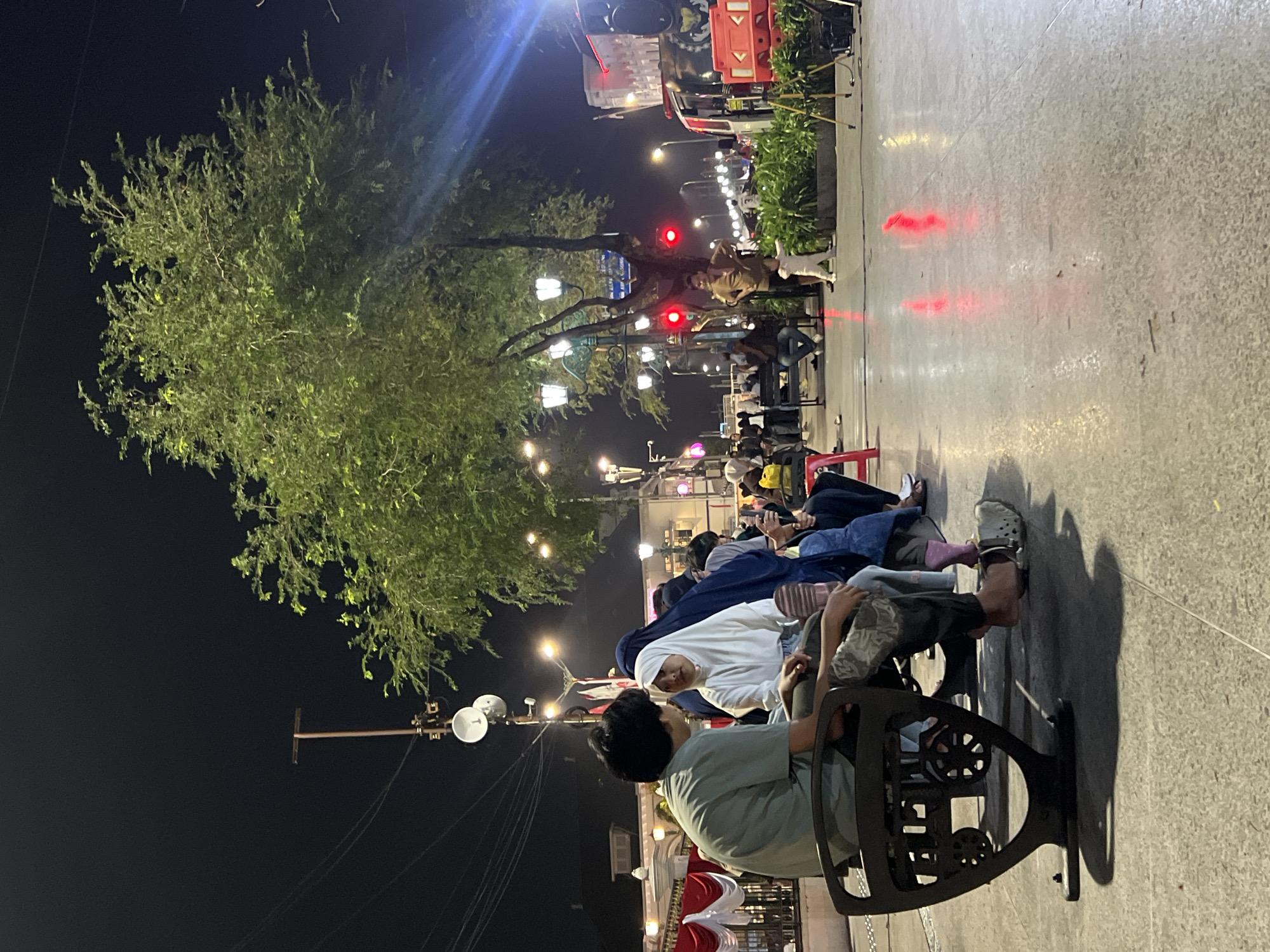 A nighttime scene captures people sitting on a dark bench in the city of Yogyakarta, Indonesia, likely along Malioboro Street, a bustling pedestrian thoroughfare known for its night markets and street vendors. The bench has an ornate, industrial design with circular elements on its base. In the foreground, a person wearing a light green shirt and patterned pants sits on the left side of the bench, with their feet resting on the ground, one wearing a white Croc-style sandal. They are engaged in conversation with another individual sitting next to them. This second person, facing the viewer, is wearing a white hijab and a light-colored top. Behind them, other people are seated on the same bench or standing, some wearing headscarves.
The background reveals a lively street scene illuminated by various lights. Overhead, a large green tree with many branches stands prominently, its leaves reflecting the ambient light. Streetlights with a classic design are visible, along with decorative string lights. A tall pole with what appear to be antennas or satellite dishes is also present. Traffic lights show red signals, indicating a pause in vehicle flow. Red and white banners and drapes are visible on buildings in the distance, adding to the festive atmosphere. The ground is paved with a light-colored, textured material that reflects the red and yellow lights from the street and surrounding establishments. The overall impression is of an active, public space at night, where people are gathered for leisure or to enjoy the city's ambiance.