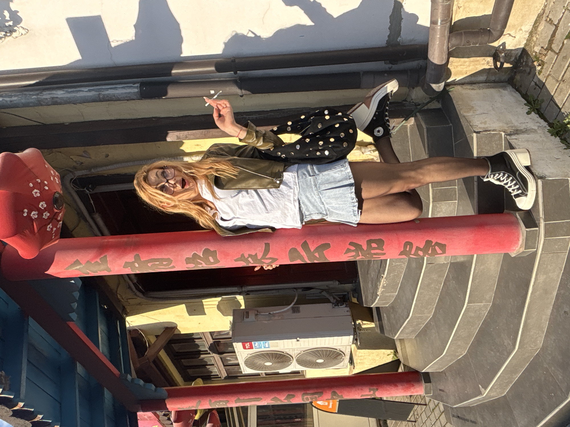A person lies horizontally across a red painted beam. The person wears glasses, blonde hair, a green jacket, a white shirt, blue shorts, and black shoes. The beam is inscribed with Chinese characters and has other beams above and below. The person is holding a cigarette in their right hand. A building with pipes and drains is in the background on one side. A set of stairs is in the background on the other side. There is an air conditioning unit visible behind the beam. The scene is outdoors in daylight. The location is not discernible as Varna, Bulgaria, from the image provided.