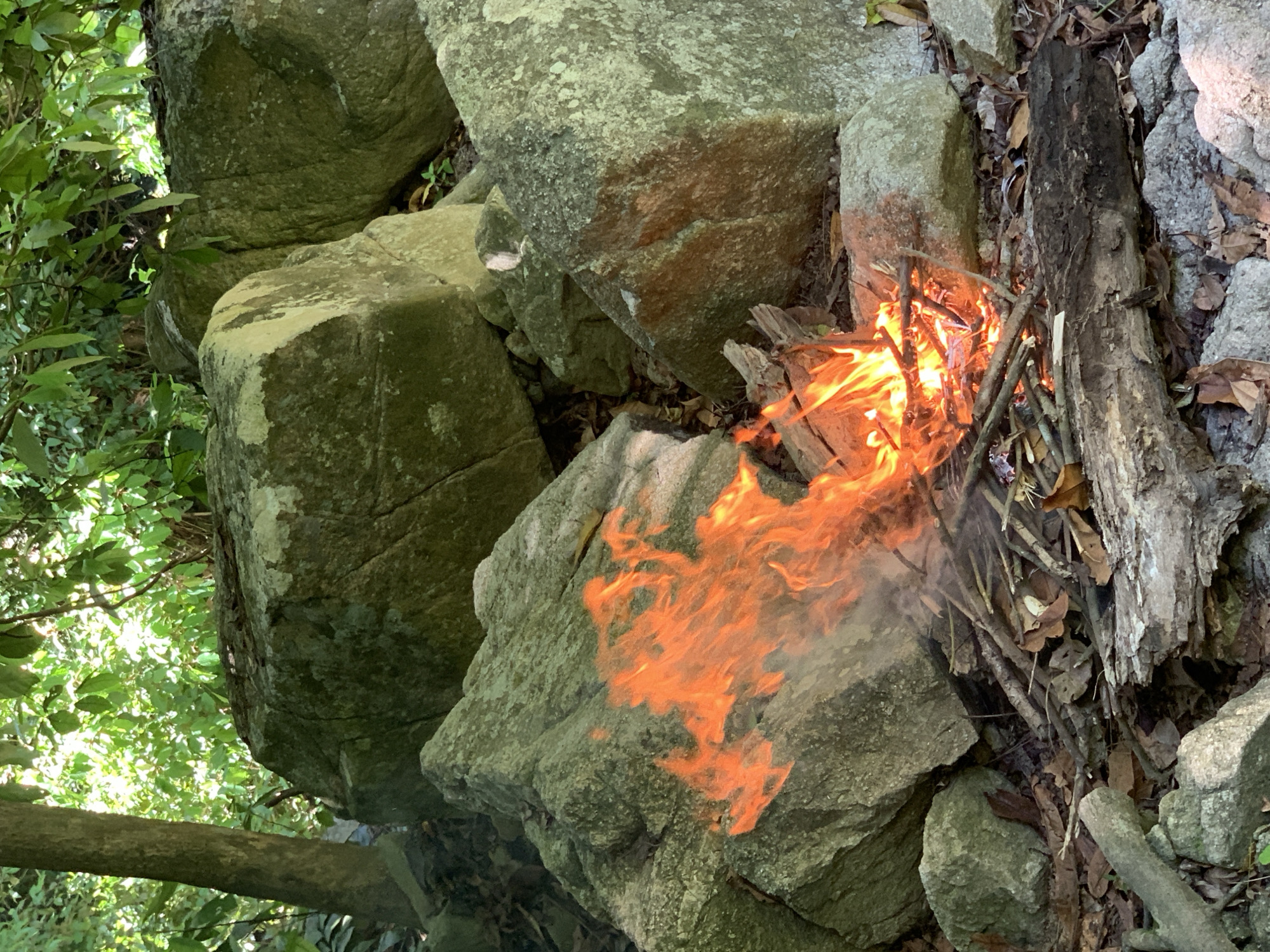 A small campfire burns in a forest clearing, surrounded by large, moss-covered rocks and fallen leaves. The fire is built with dry twigs and branches, and the flames are a vibrant orange and yellow. Sunlight filters through the dense green foliage overhead, casting dappled shadows on the ground. There are no people or animals visible in the image. The location appears to be a natural, outdoor setting, possibly a hiking trail or picnic area within the Ko Samui District of Thailand.