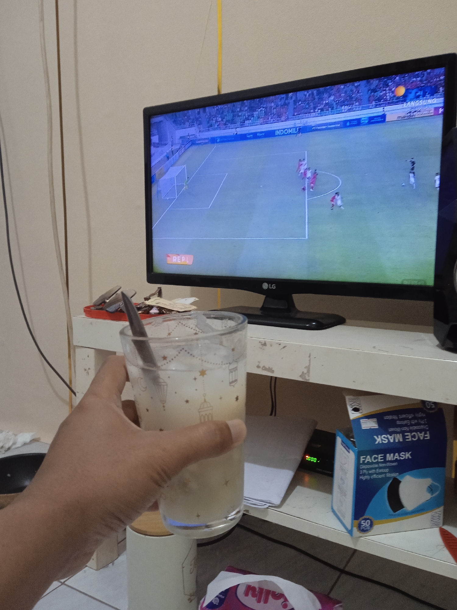 This image captures an indoor scene, likely a residence in Brangsong, Indonesia, where a person is engaged in leisure activities. The foreground shows a light-skinned hand holding a clear glass containing a light, opaque beverage with ice and a metal spoon. The glass is adorned with patterns resembling stars and Islamic lanterns. In the midground, a black LG television displays a soccer match in progress, featuring players in red and white jerseys on a green field with a white goal. Overlays on the screen include "INDOMILK," "LANGSUNG," and "REPL." The television rests on a white, weathered wooden shelf. To the left of the TV on the shelf, a red tray holds various metal clips and small items. Below the television, on a lower shelf, a stack of white papers, a small black device with green digital numbers (likely a clock or timer), and an open blue and white box labeled "FACE MASK" (indicating 50 disposable 3-ply masks) are visible. A black speaker is partially seen to the right of the TV. The background consists of a plain, light-colored wall with vertical panel lines and various black electrical wires running along it. A yellow cable extends vertically in the upper right. On the floor or a lower surface, a white ceramic container with a wooden lid and part of a pink and white plastic bag are visible.