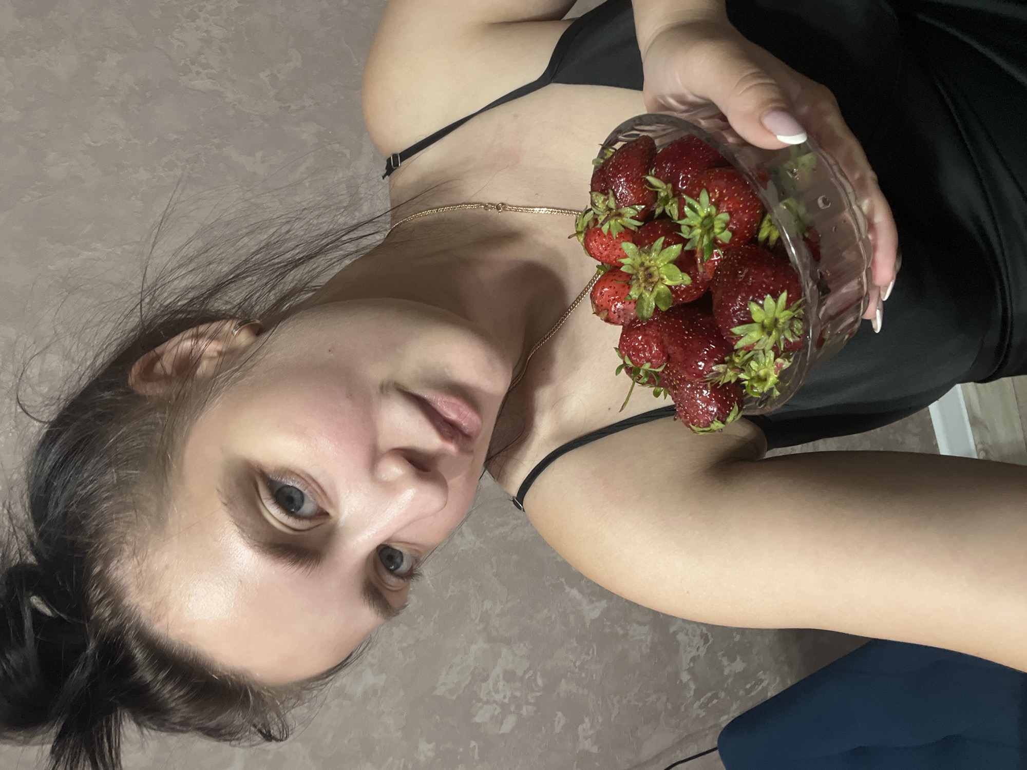 The image shows a person holding a bowl of strawberries. The person is wearing a black top and a gold necklace. The background is a textured surface, possibly a wall or floor. The strawberries are red and appear fresh. The lighting suggests it might be daytime, but there are no strong indicators of time or weather. The location is noted as Марковский, Russia. The image is taken from a close-up perspective, focusing on the person and the strawberries. The person's face is partially visible, with their eyes looking towards the camera. The overall mood is casual and relaxed. There is no visible text in the image.
