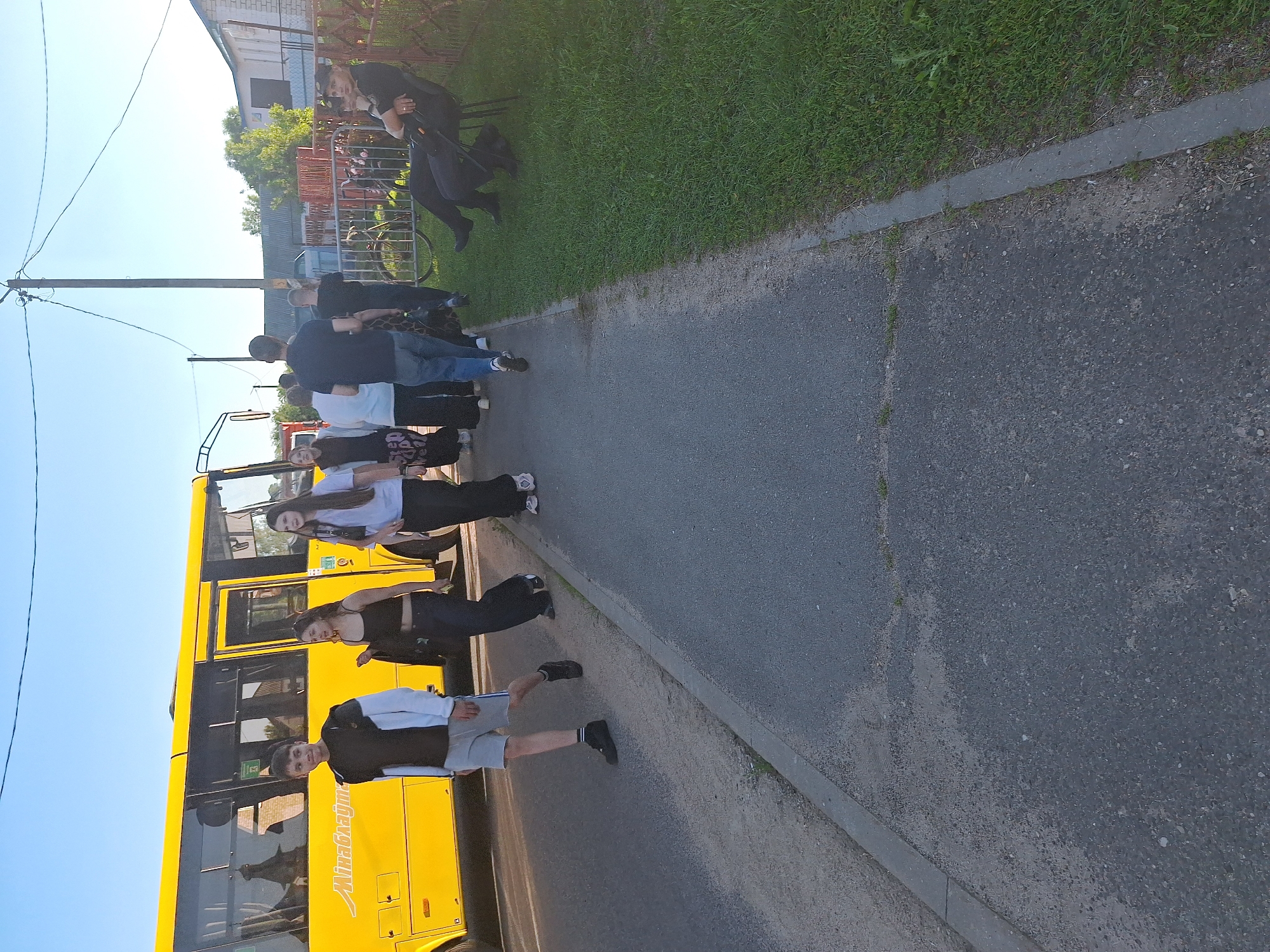 On a paved path next to a road, a group of people are walking. On the left, a bright yellow bus is parked. A young man in a black and white jacket and grey shorts is walking towards the camera. To his right, a young woman in a black tank top and dark pants is walking. Another young woman in a white t-shirt and black pants is walking behind them. Further back, several other individuals are walking away from the camera, including a man in a dark t-shirt and jeans. To the right of the path, a grassy area is visible, with a fence and a building in the background. Two people are sitting on chairs in the grassy area, one of whom appears to be wearing a dark uniform and a cap. The sky is clear and blue. The scene appears to be in a residential area.