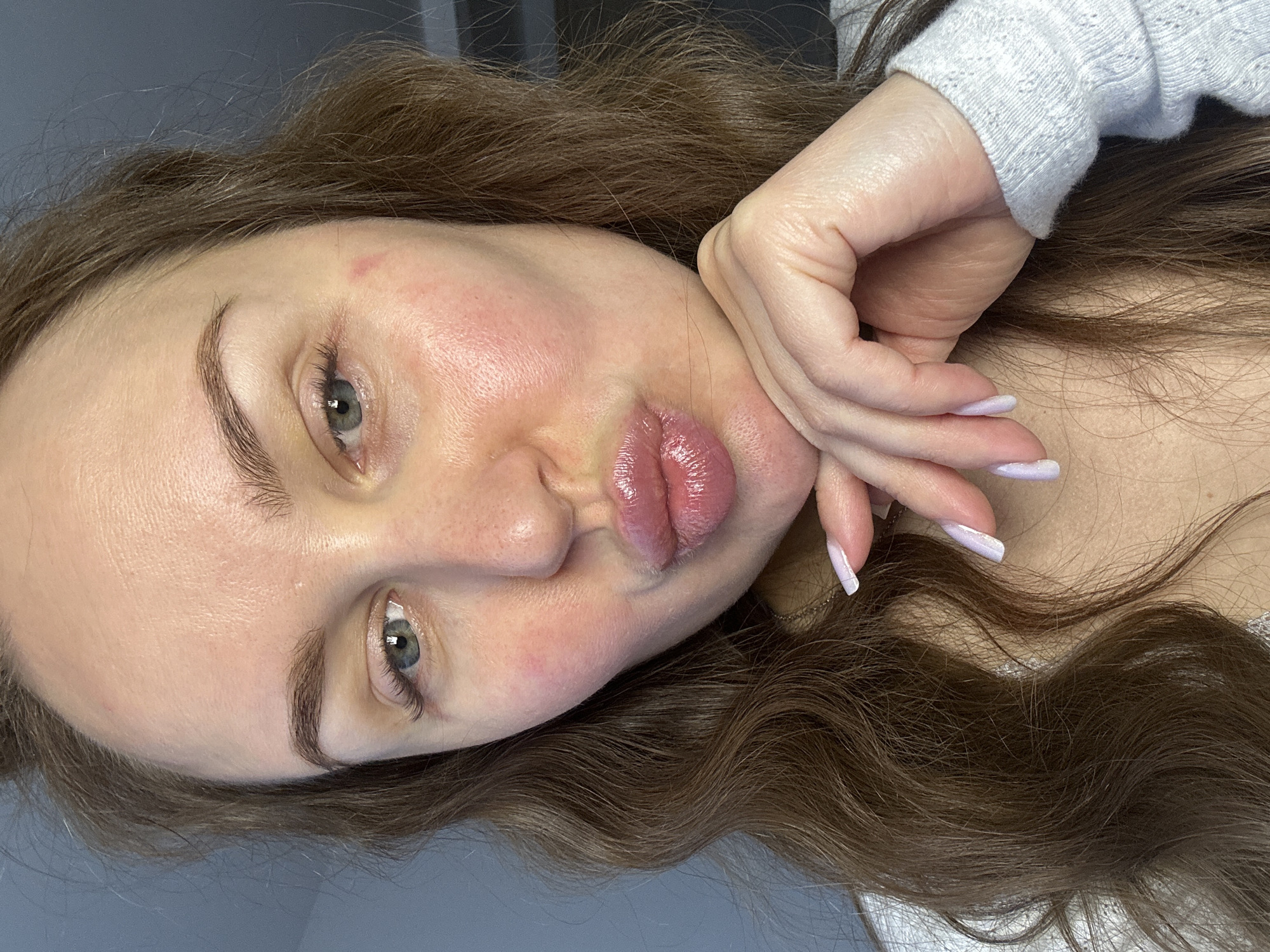 An adult female subject is depicted in a close-up portrait, located in the city of Budva, Montenegro. She has long, wavy brown hair, blue-grey eyes, and fair skin with localized redness on her cheeks and forehead. A distinct linear red mark is present on the subject's right cheek. Her lips are pursed in a 'kiss' gesture and appear glossy. Her right hand, with long, light lavender-painted nails, supports her chin. She wears a light-colored, textured knit garment, possibly light blue or grey, and a thin, gold-colored chain is partially visible around her neck. The background consists of an out-of-focus, plain, light blue-grey wall or surface. The subject is looking directly into the camera.
