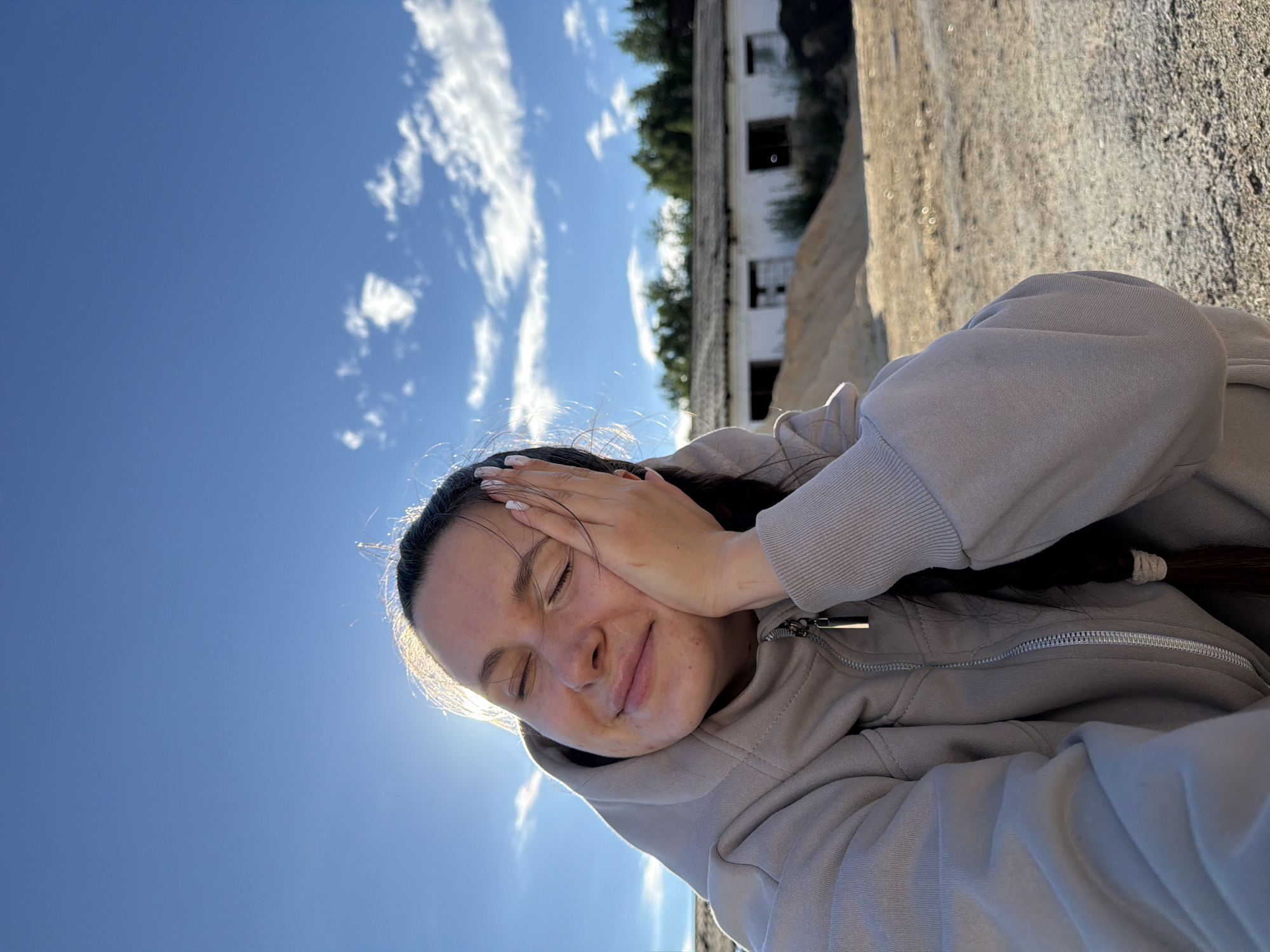 A young woman with her eyes closed is resting her cheek on her hand, smiling serenely. She is wearing a light gray zippered hoodie. Her dark hair is braided into a single plait that rests over her shoulder. The background shows a bright blue sky with scattered white clouds. In the distance, there is a long, low-slung building with a dilapidated appearance, partially obscured by trees. The ground in front of the building is a mixture of dirt and sand. The overall setting appears to be an outdoor, possibly rural or industrial area. The image is taken outdoors on a sunny day. The location is Красный Маяк, Russia.
