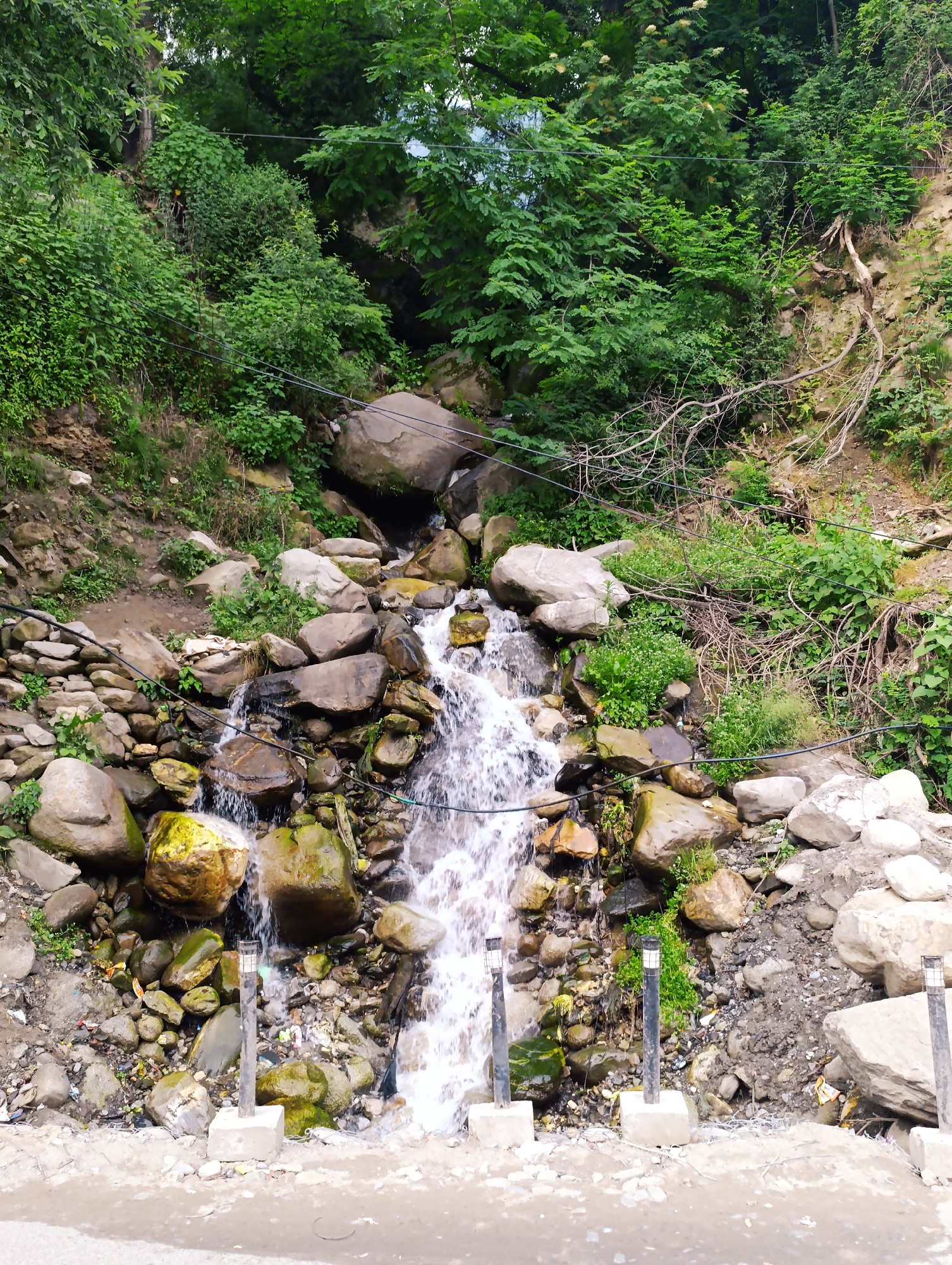 The media file depicts a natural scene in Vashisht, India. A waterfall cascades down a rocky slope, surrounded by lush green vegetation and large boulders. The water flows over a jumble of stones, creating a dynamic and frothy effect. Several modern, short lamp posts are visible in the foreground, suggesting the presence of a pathway or roadside. The overall impression is one of a natural landscape enhanced by man-made elements. The time of day appears to be daytime, with soft, diffused light indicating potentially overcast or cloudy weather, typical for the monsoon season often experienced in hilly regions of India. The presence of greenery suggests a favorable climate for plant growth. The image conveys a sense of natural beauty and tranquility, with a touch of ruggedness from the rocks and slope. There is no visible text in the media file.