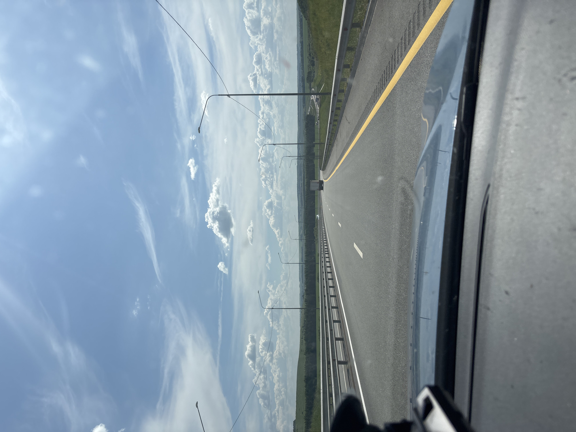The image depicts a view from inside a vehicle traveling on a multi-lane highway under a partly cloudy sky. The highway has a yellow line marking the right edge of the lane and a white dashed line separating lanes. Metal guardrails run along the side of the road. Several streetlights are visible along the highway. The surrounding landscape consists of green, rolling hills and a treeline in the distance, suggesting a rural or semi-rural environment. A white truck is visible in the distance on the highway. The interior of the vehicle, including the dashboard and a mount for an electronic device, is in the foreground. The image is taken from a first-person perspective, looking forward along the road. There is no information in the image to confirm its location in Старое Русское Маматкозино, Russia.