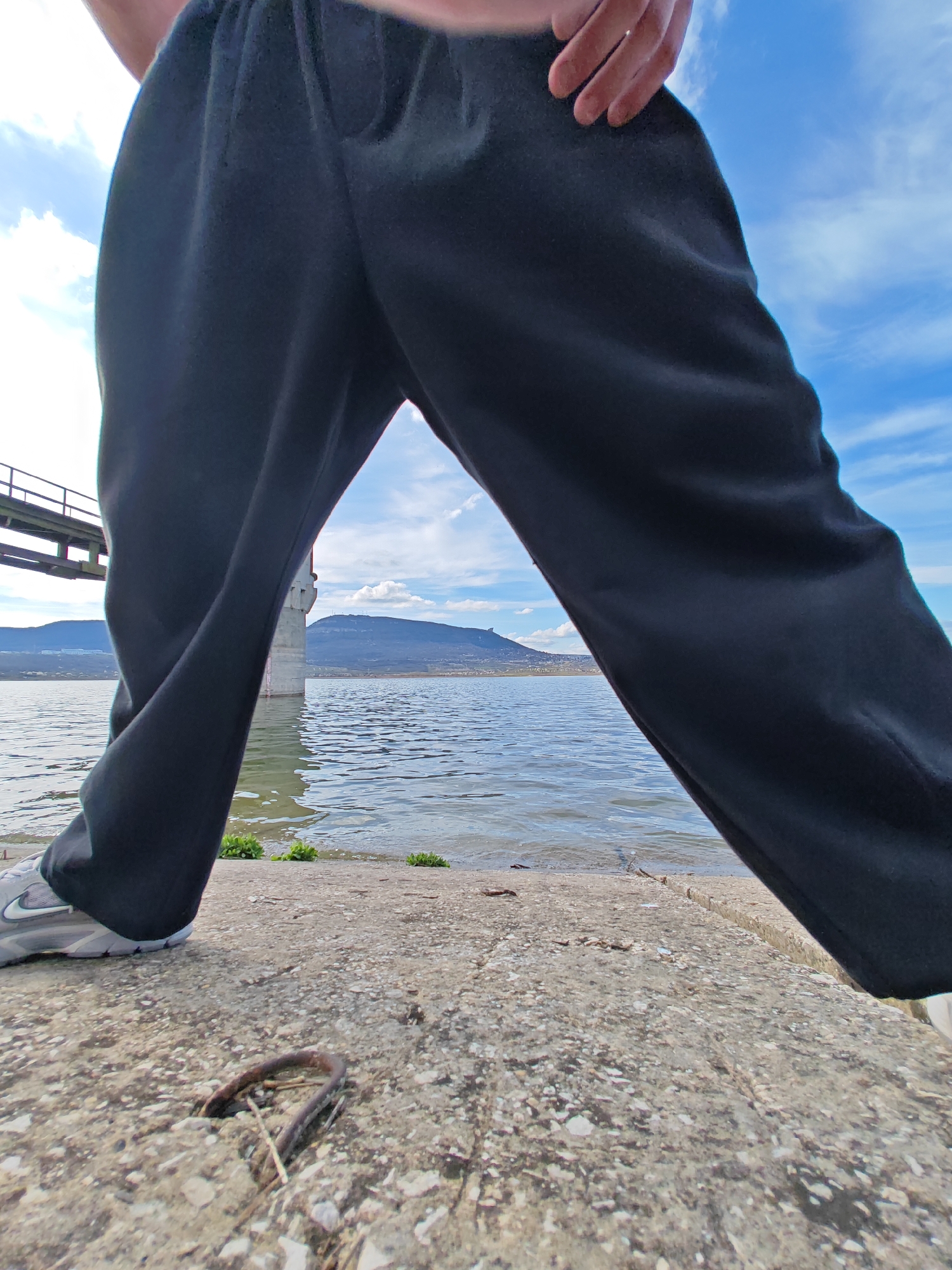 The image presents a ground-level, wide-angle perspective of a person standing on a concrete ledge near a body of water. The person is wearing dark-colored sweatpants and grey sneakers with a white swoosh logo. A hand is visible near the waistline. The foreground features a textured concrete surface with a protruding metal loop. Beyond the ledge is water with small patches of vegetation along the shoreline. In the background, there is a bridge pier and a mountain range under a partly cloudy sky. The location appears to be near the Shumen lake, Bulgaria.