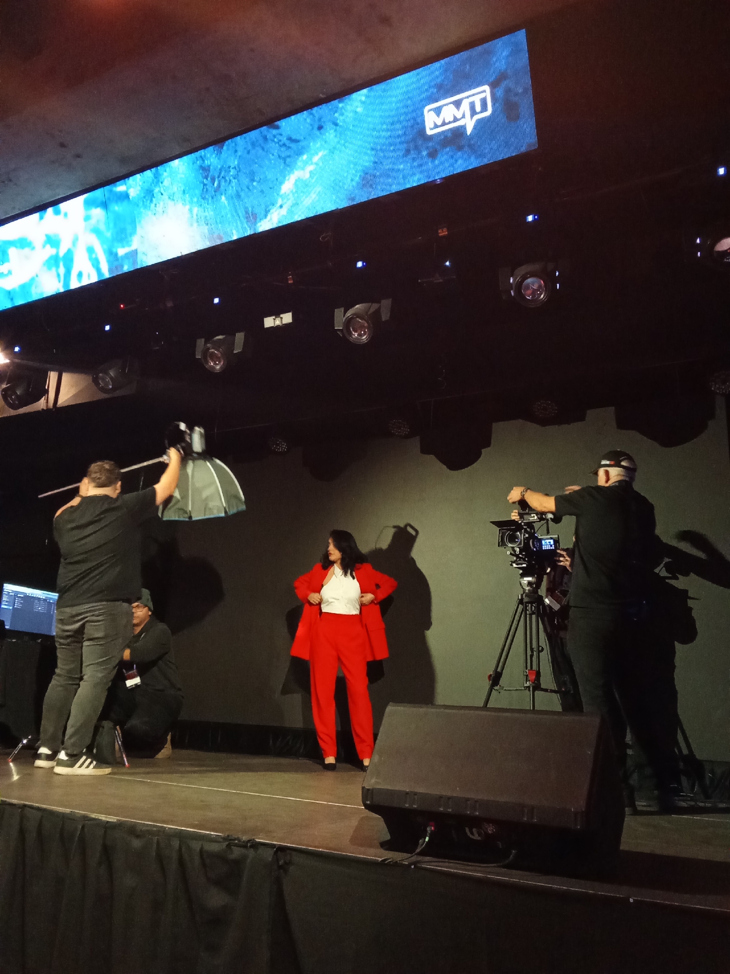 A woman in a vibrant red suit and white top stands center stage, posing as a photographer on the left adjusts a boom mic. To the right, a cameraman operates a professional video camera on a tripod. Another person kneels on the stage near the photographer, assisting with equipment. The setting is a dimly lit indoor venue, possibly a conference or presentation space, with a large screen displaying a blue, abstract image and the "MMT" logo at the top. Stage lighting is visible above, casting shadows on the dark backdrop. The overall atmosphere suggests a professional media production or event in Santiago, Chile.