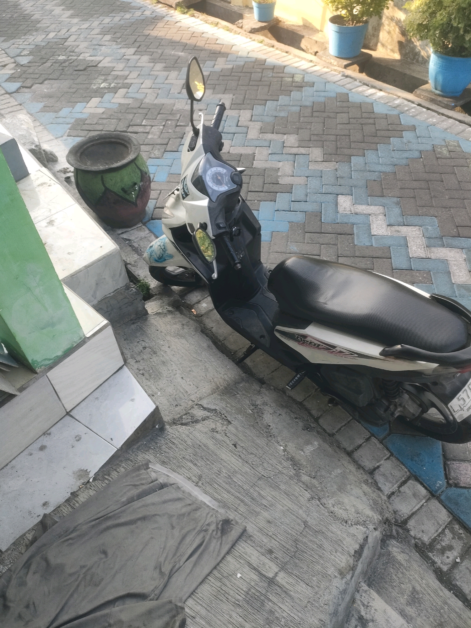 A white and black scooter is parked on a tiled walkway. The walkway is composed of rectangular tiles arranged in a herringbone pattern, with alternating sections of gray, light blue, and white. To the left of the scooter, there is a large, green and red patterned pot. Next to the pot, a concrete structure with green and white tiled sections rises. In the foreground, a gray piece of cloth is draped over a concrete surface. In the background, several blue potted plants are visible, placed on a raised concrete edge. The scene appears to be an outdoor public space, possibly a sidewalk or courtyard, with the characteristic urban environment of Surabaya, Indonesia.