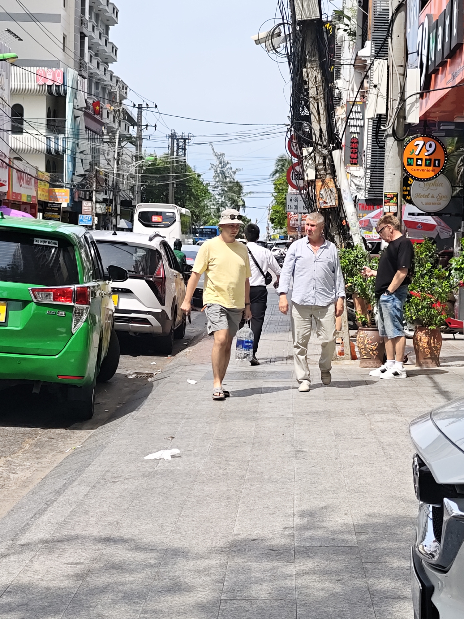 A group of people are walking along a sidewalk in what appears to be a busy street in Vietnam. The scene is characterized by a dense network of electrical wires hanging overhead, typical of urban areas in Southeast Asia. Two men are prominently featured in the foreground, walking side-by-side. The man on the left wears a yellow t-shirt and grey shorts, while the man on the right is dressed in a light blue button-down shirt and light-colored pants. Behind them, another man in a black t-shirt and denim shorts is visible, looking at his phone.
In the background, various vehicles are parked along the street, including a bright green car on the left and a white car behind it. A white bus is also visible further down the street. The buildings lining the street are multi-story structures, some with balconies and awnings. Signs with text in what appears to be Vietnamese and English are displayed on the buildings and poles, advertising businesses such as "AZURA HOTEL," "Sophia Hotel & Spa," and "79 Mart Convenient." One sign also indicates store hours from "6:30-16:30-18." The weather appears to be sunny, casting shadows on the sidewalk, suggesting it is daytime. The overall atmosphere is that of a bustling urban environment.