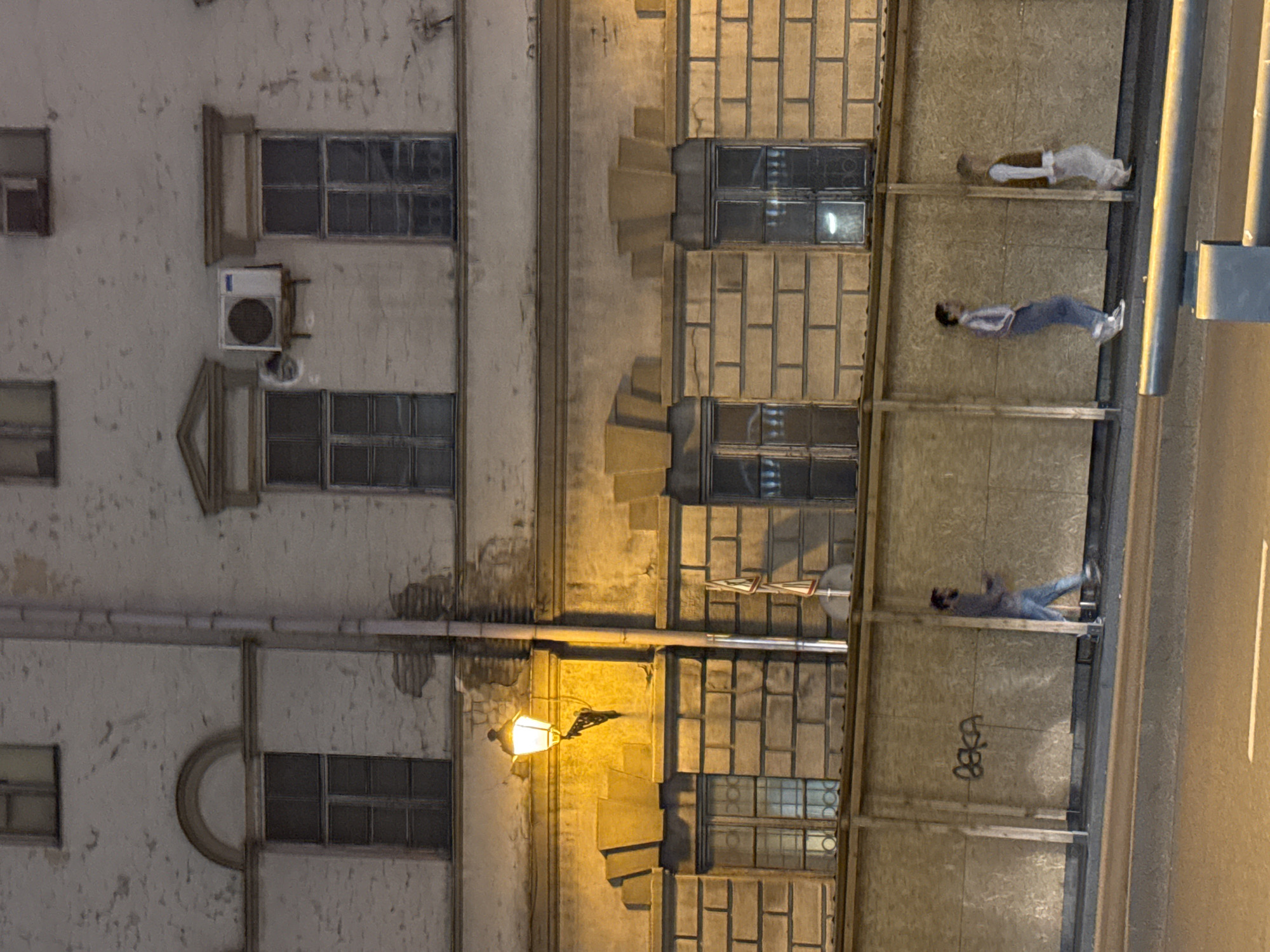 The image shows a building facade and what appears to be an elevated walkway in Tbilisi, Georgia. The building is multi-story, with windows visible on the left side. An air conditioning unit is mounted on the facade. A street lamp is attached to the building.
On the right, an elevated walkway or structure with a textured wall is visible. Several people are present on this walkway, either lying down or in a reclined position. There are at least three individuals visible. A small amount of graffiti is also seen on the wall. A road with a light appears on the right. The photo seems to be taken at night or in low light.