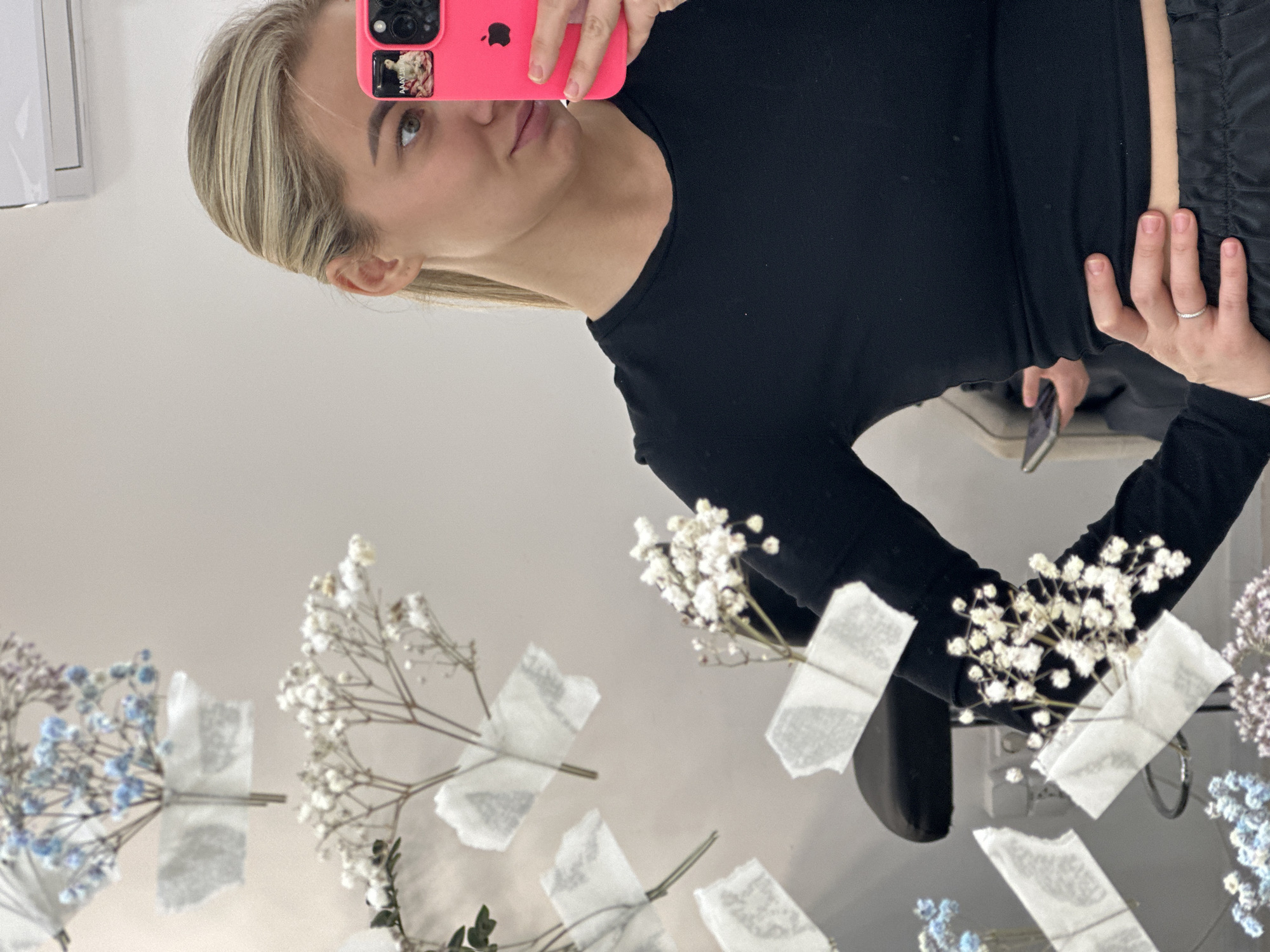 This image captures a mirror selfie taken by a young woman in an indoor setting, presented with a 90-degree clockwise rotation. The subject is a woman with blonde hair and light-colored eyes, wearing a black long-sleeved top. She is holding a bright pink smartphone with a visible Apple logo and multiple camera lenses in her right hand. A small picture or sticker with faint, mostly illegible text, possibly "MAYYLEN," is visible on the phone case near the camera. Her left hand rests on her waist, displaying a thin, stone-studded ring on her ring finger.
The selfie is taken in a reflective surface, likely a mirror or a highly polished table, which is adorned with several small bouquets of baby's breath flowers. These delicate sprigs, featuring white, light blue, and light purple blossoms, are secured to the reflective surface with pieces of white, semi-transparent tape, creating a decorative effect. In the reflection, another smartphone is partially visible in the background, suggesting another person or object within the space.
The background behind the woman appears to be a plain, light-colored wall, contributing to a clean and bright aesthetic. The woman's expression is neutral as she looks directly into the reflected phone lens. The setting suggests a stylized interior, possibly within Budva, Montenegro, providing a unique backdrop for the selfie.