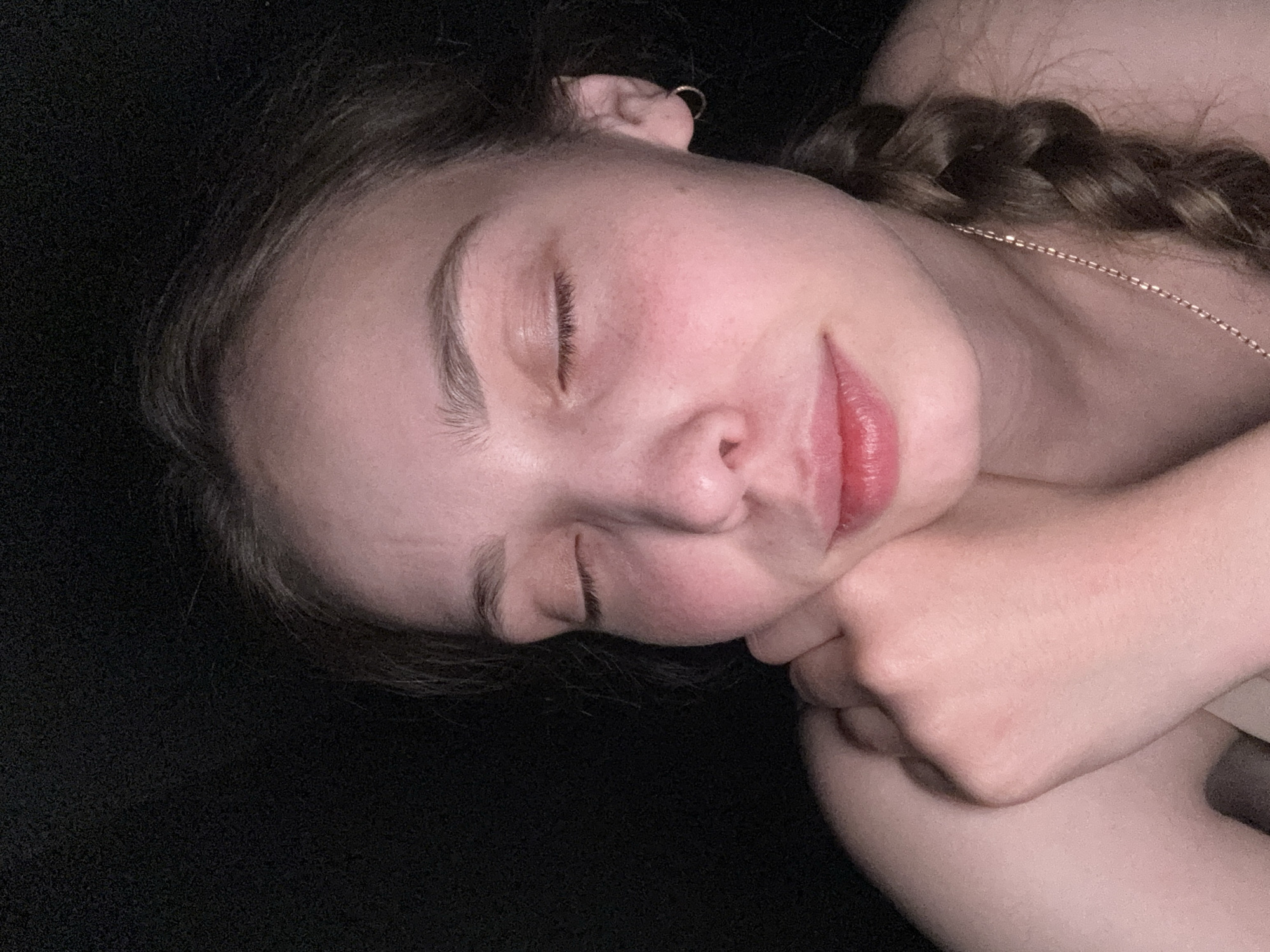 A close-up, diagonal portrait captures a young person, likely female, resting or sleeping peacefully against a dark background. Her eyes are closed, and her head is turned to the right, with her hands gently folded or supporting her face. She has light skin, visible faint blush on her cheeks, and dark brown hair styled in a braid that is partially visible on the right side of the frame. A small golden hoop earring adorns her left ear, and a delicate golden chain is seen around her neck. Her lips are a natural reddish-pink color. The dark background, possibly a blanket or fabric, suggests an indoor, intimate setting, perhaps a bedroom, and implies the time of day is either night or a darkened room, conducive to sleep or rest. The overall mood is calm and serene. No text is visible in the image.