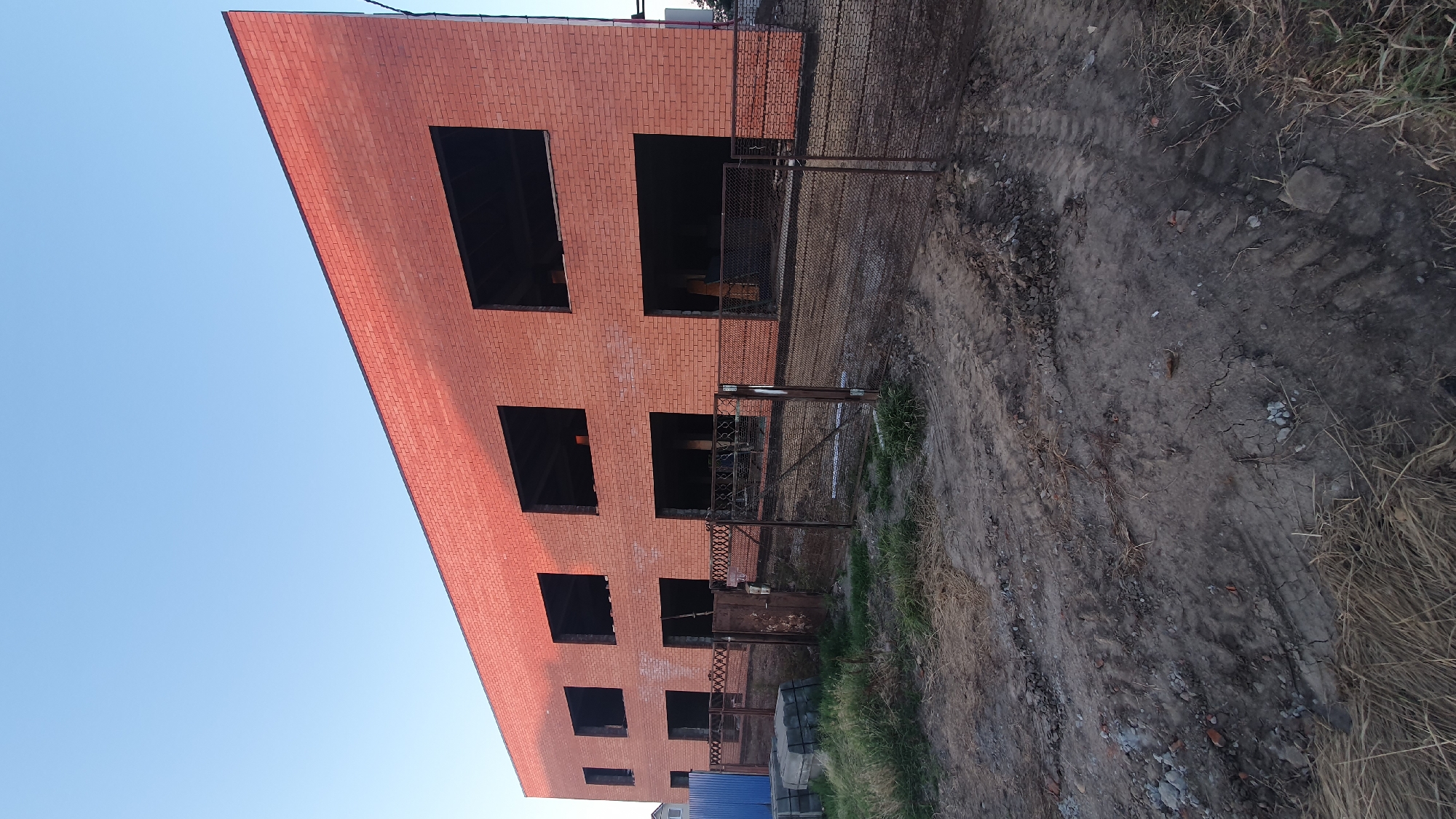 The image depicts the construction of a red brick building in Ust-Labinsk, Russia. The building has two stories with multiple large, dark openings where windows and doors will be installed. A metal fence encloses the construction site, and piles of building materials, including concrete blocks and bricks, are visible. The ground is rough and unpaved, with tire tracks suggesting recent vehicle activity. The sky above is clear and blue. There are no people visible in the image.
