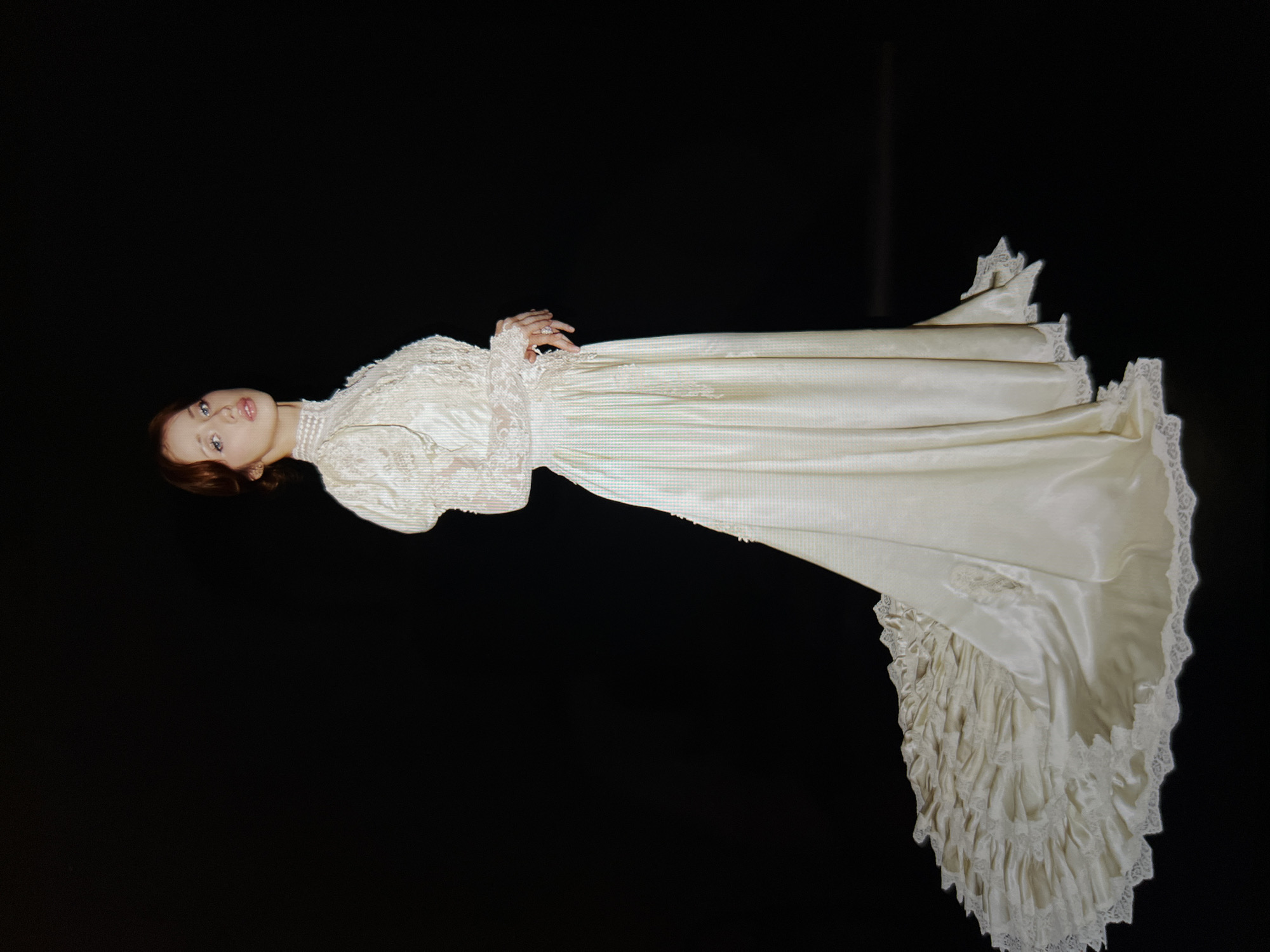A young woman with auburn hair styled in an updo and light makeup stands facing the viewer and looking slightly to her right. She is wearing a vintage-style white wedding dress with a high lace collar, puffed sleeves, and long lace sleeves. The dress has a fitted bodice and a voluminous skirt that extends into a long train adorned with ruffles and lace trim. Her hands are clasped in front of her, and she wears rings on her fingers. The background is entirely black, creating a studio portrait effect. There are no discernible elements that would indicate a specific location within a city or country.