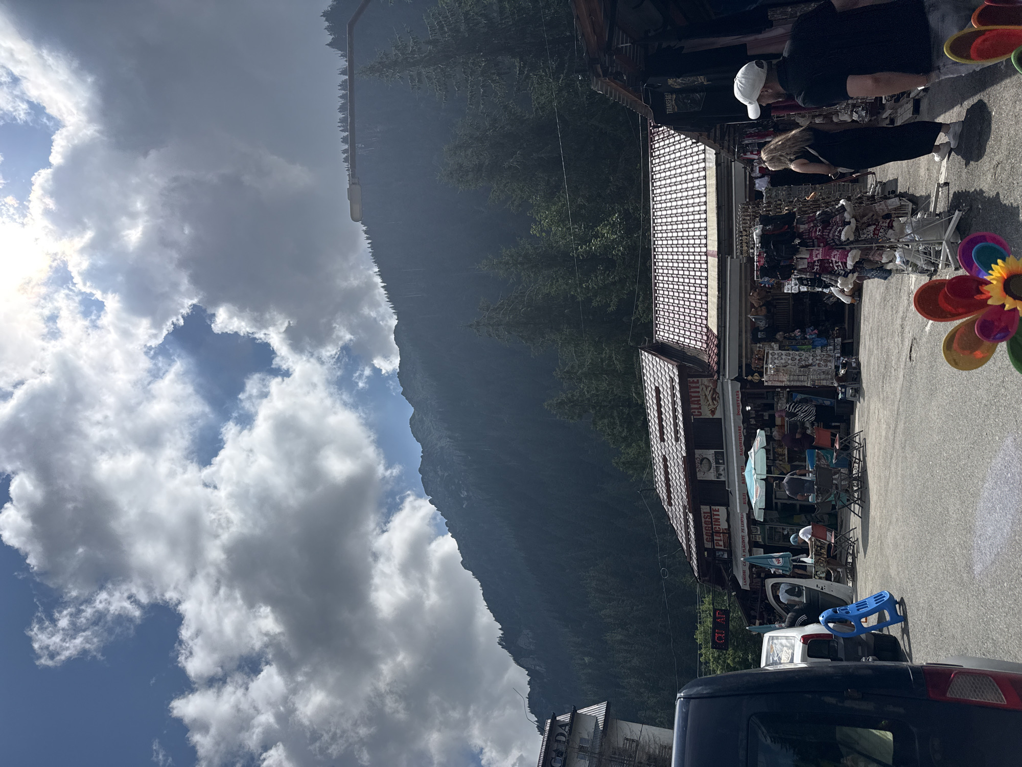 In Cârțișoara, Romania, a mountain landscape dominates the background with a dark, heavily forested slope rising towards a jagged peak. The sky above is a mix of bright blue and dramatic, voluminous white and gray clouds, with sunlight piercing through in some areas. In the foreground, a street scene unfolds with several buildings, possibly shops or stalls, featuring red tiled roofs. These structures appear to be selling various goods, with racks of clothing and other items displayed outside. Several people are visible: a man in a black shirt and white cap walks away from the camera, accompanied by a woman in a black dress. Others are seated at tables under umbrellas near one of the establishments. A dark-colored van is parked on the left side of the frame, and a bright blue plastic object, possibly a child's toy or a piece of equipment, is propped against it. Colorful pinwheels are placed in the foreground, adding a touch of vibrancy to the scene. The overall impression is a busy outdoor market or rest stop situated at the foot of a majestic mountain range.