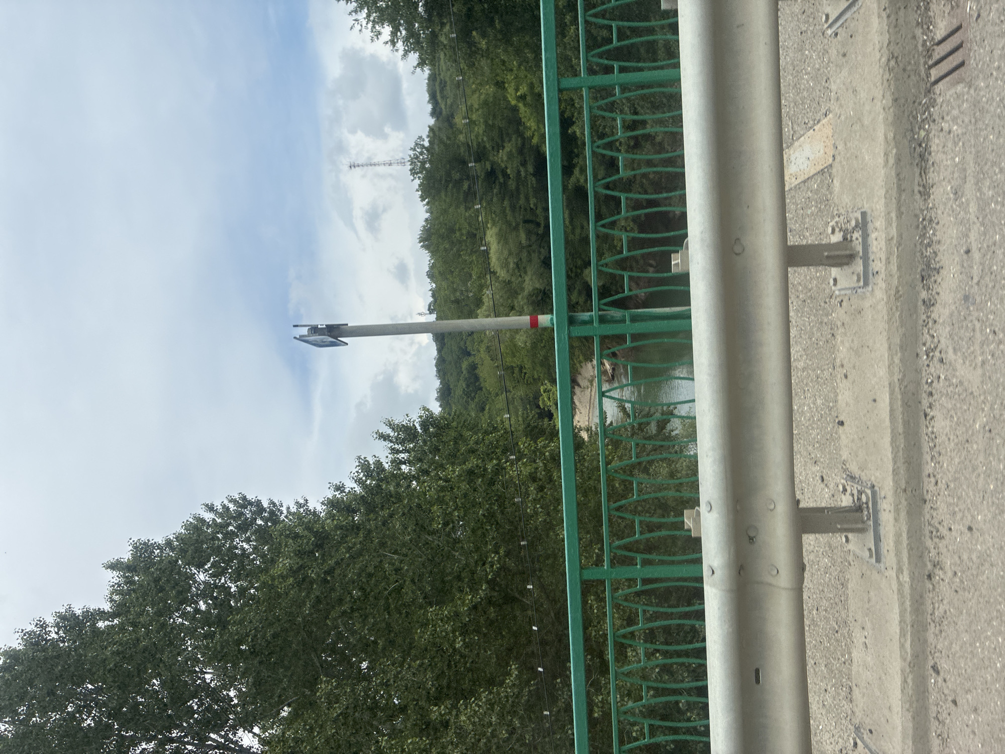 The image shows a view from a bridge in Arkhipo-Osipovka, Russia. A green railing with a decorative pattern is visible in the foreground, with a silver guardrail above it. Beyond the railing, a body of water, likely a river, can be seen flowing through a wooded valley. Tall trees with dense green foliage line the banks of the river and cover the rolling hills in the background. A tall metal tower, possibly a communication mast, is visible on one of the hills. The sky is overcast with white clouds. A lamppost with a sign is positioned on the bridge. The road surface of the bridge is visible at the bottom of the image.