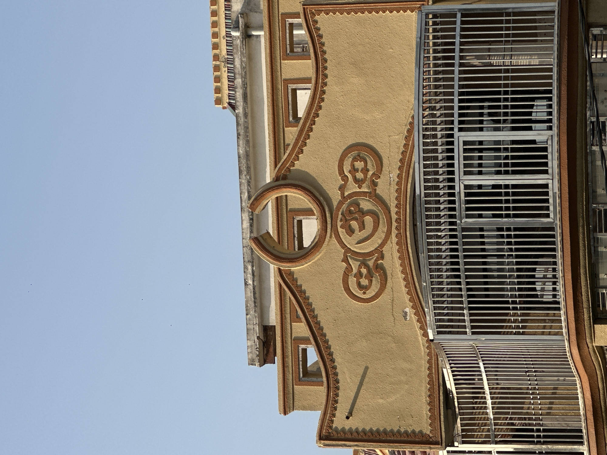 A low-angle, eye-level shot of a building facade in Mathura, India. The building is painted beige with brown trim and decorative elements. A prominent circular ornament containing the Om symbol is visible. A metal-grilled balcony is attached to the building's side. The sky is a clear light blue.