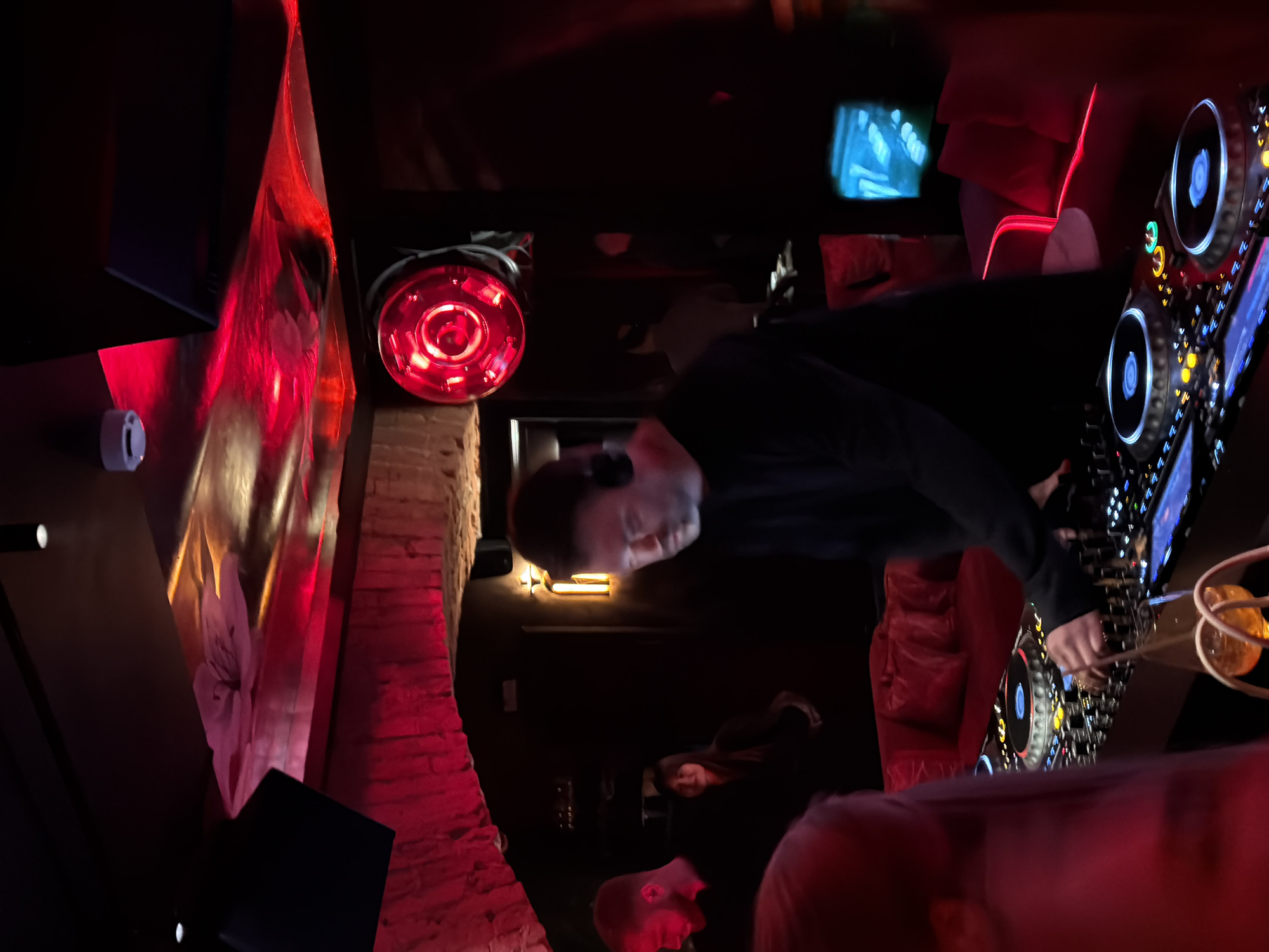 A DJ is shown mid-performance, possibly in a nightclub or bar. The scene is dominated by red lighting, illuminating a brick wall on the left and an artistic installation of what appears to be draping fabric. The DJ is wearing headphones and a dark shirt, focused on the DJ equipment in front of him. Other people can be seen blurred in the background and foreground. The equipment features illuminated buttons and dials. The image was taken in a dark environment. It is not possible to confirm the location in Moscow, Russia.