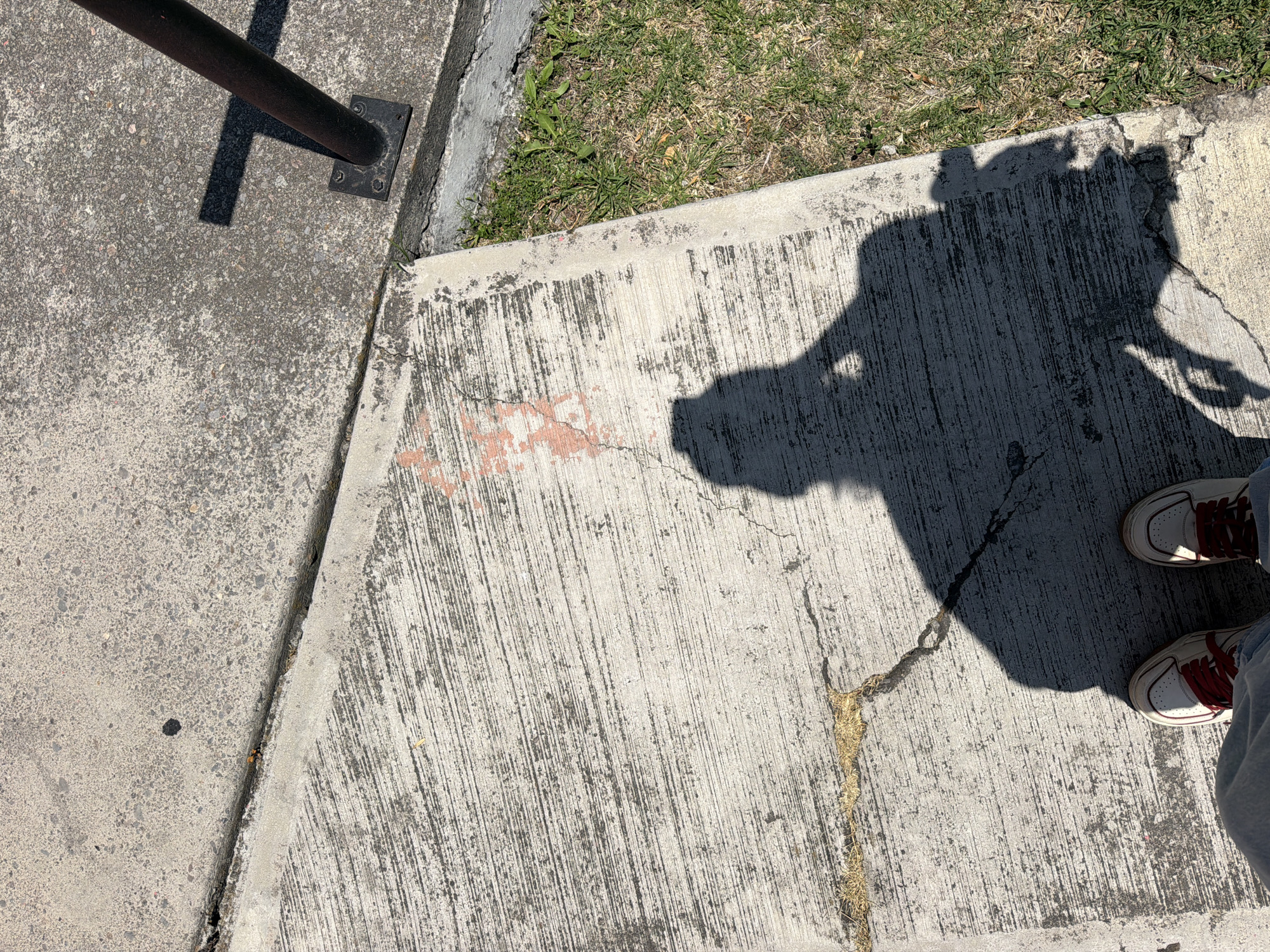 The image shows a section of concrete sidewalk in San Andrés Cholula, Mexico. A metal bollard is visible on the left side. The shadow of a person is cast onto the sidewalk, including shoes and a hand. There is grass next to the sidewalk. The concrete is cracked in places and has a pinkish stain in the center.