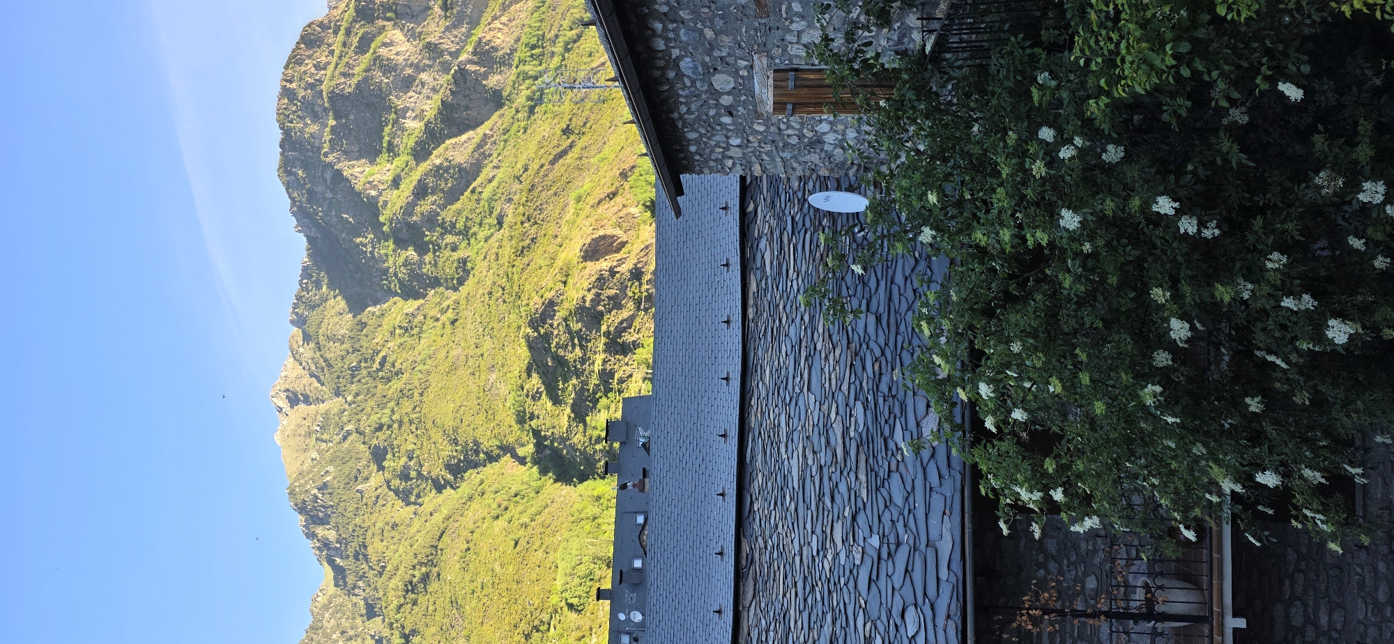 The image captures a view of the Pyrenees mountains in Benasque, Spain, alongside a building with a slate roof and stone wall. A lush bush with white flowers grows near the wall. The sky is blue. The mountain is covered with green vegetation. A satellite dish is mounted on the roof.