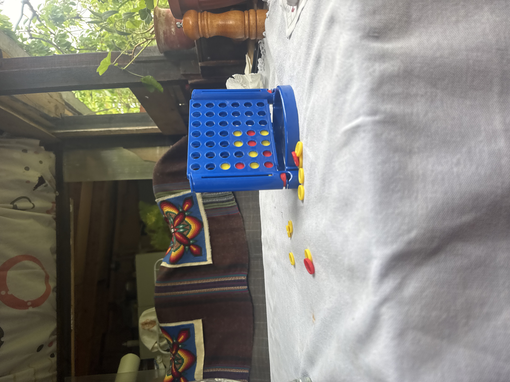 The image shows a partially filled blue Connect Four game board on a white tablecloth. Several yellow and red game pieces are scattered beside the board. The scene appears to be indoors, possibly in a sunroom or enclosed porch based on the presence of exterior views through a window. The location and city cannot be determined from the image provided.