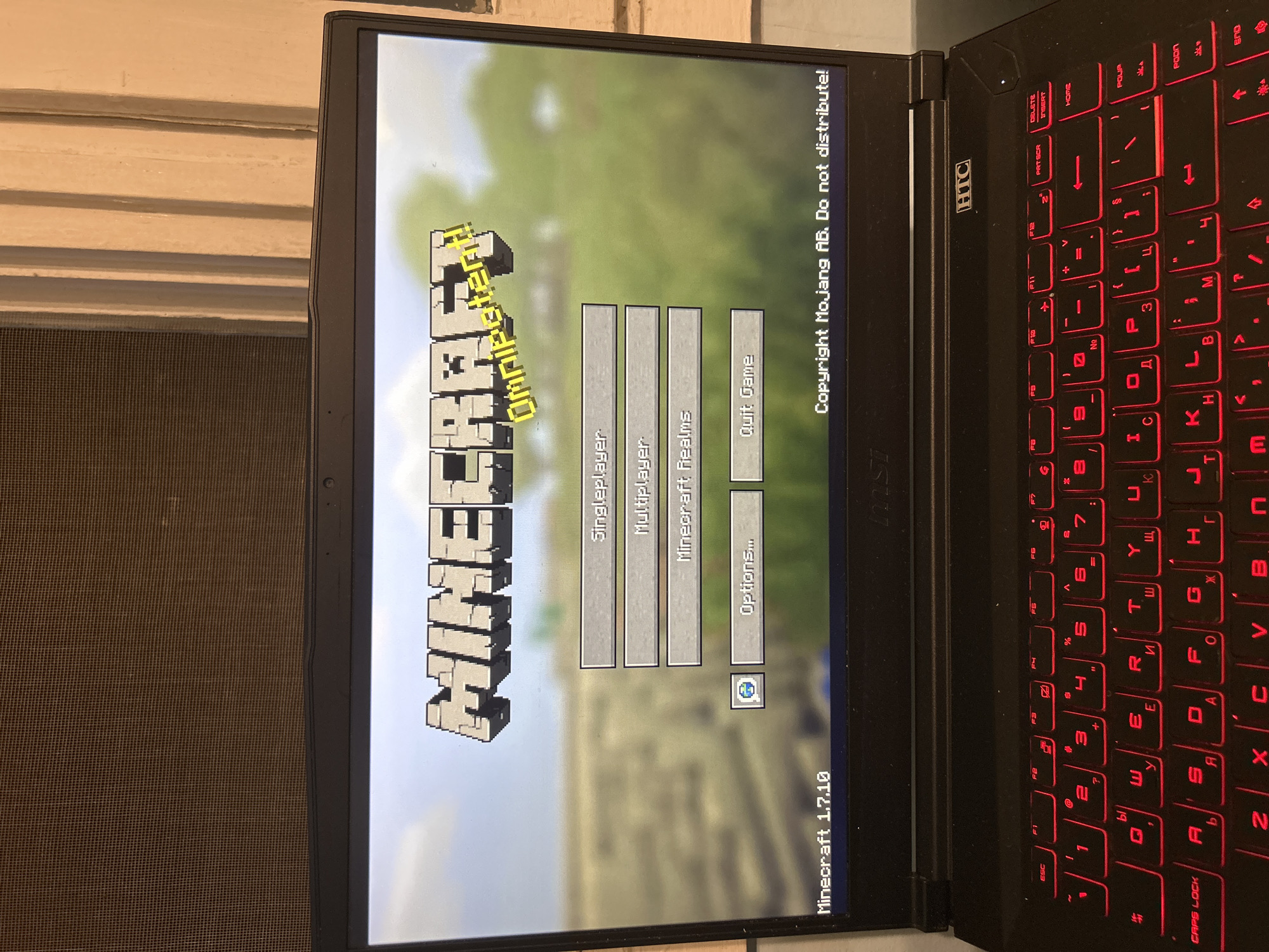 The image shows a laptop screen displaying the main menu of the video game Minecraft, version 1.7.10. The menu options are "Singleplayer", "Multiplayer", "Minecraft Realms", "Options...", and "Quit Game". The copyright notice "Copyright Mojang AB. Do not distribute!" is visible. The laptop has a red-backlit keyboard with some Cyrillic characters. The make of the laptop is MSI. The laptop appears to be sitting next to a window.