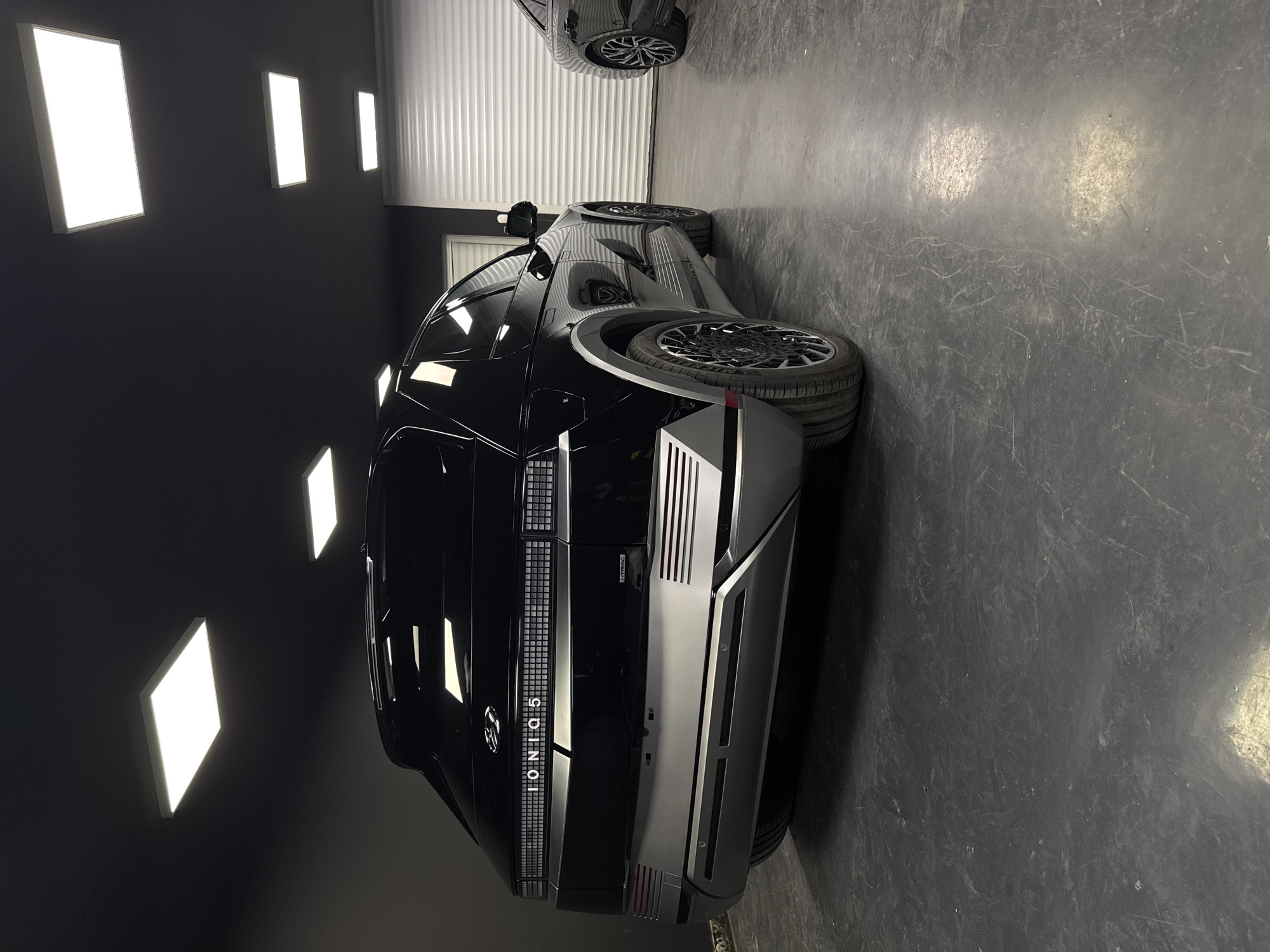 The image shows the rear passenger side view of a black Hyundai Ioniq 5 electric vehicle parked in a garage. The vehicle's taillights are illuminated, and the "IONIQ 5" badging is visible. The garage has a dark ceiling with recessed lighting panels. The floor is dark gray concrete with some scuff marks. To the right of the Ioniq 5, another dark-colored vehicle is partially visible. The overall environment suggests a modern, well-lit automotive display or service area. There are no people visible in the image.