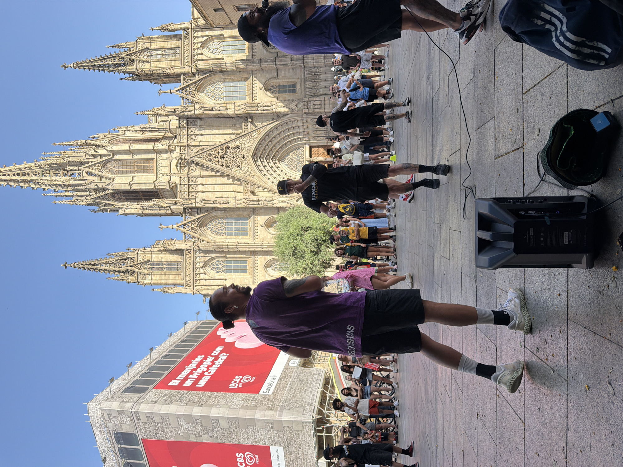 The image depicts an outdoor scene in Barcelona, Spain, with the Barcelona Cathedral prominently featured in the background. In the foreground, a group of people are gathered, appearing to be part of a performance or event. One man in a purple shirt and black shorts is facing away from the camera, holding a water bottle. Another man in a black t-shirt and shorts, wearing a black cap, is facing the camera and appears to be speaking or singing into a microphone. A third man, also in black attire, is standing behind the man in the black t-shirt. Several other individuals are standing around, some watching the performance.
The environment is a paved plaza, with a large billboard in the background featuring red and white lettering and an image of a pink ice cream. The weather is clear and sunny, with a bright blue sky. There is a black speaker on the ground in the foreground, with a cable running from it. A black bag with a green interior and a blue object is also on the ground near the speaker. The architecture of the Barcelona Cathedral, with its Gothic spires and intricate stonework, dominates the background, indicating the specific location within the city.