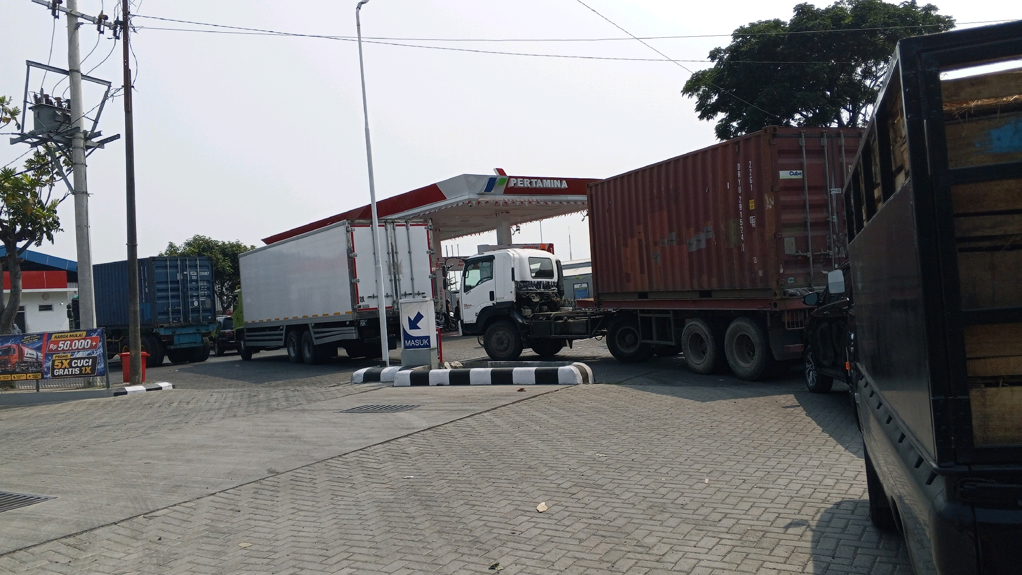 The image shows a Pertamina gas station in Semarang, Indonesia, with several large trucks parked. The gas station features a prominent red and white canopy with the Pertamina logo. A white box truck is parked directly under the canopy, with a dark red shipping container truck to its right. To the left of the white box truck, a blue shipping container truck is visible. A black truck with a wooden cargo bed is seen in the foreground, partially obscuring the view of the other vehicles. The ground is paved with interlocking bricks. A traffic sign with an arrow pointing left and the word "MASUK" (Indonesian for "enter") is positioned near the entrance. A billboard advertising prices and promotions for fuel and car washes is located on the left side of the image. Power lines and a utility pole are also present in the scene. The sky is overcast.