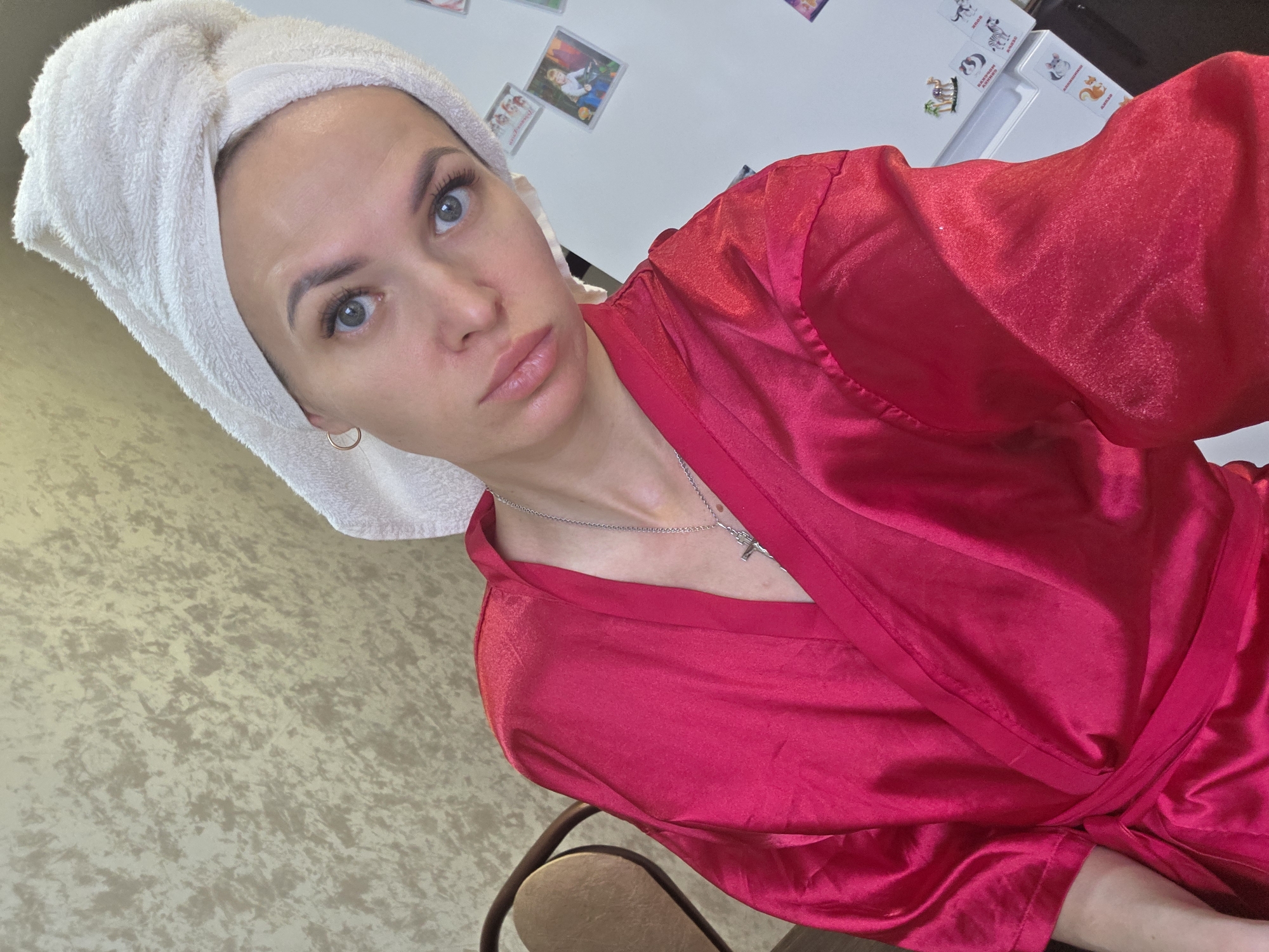 A woman is posing for a selfie, wearing a white towel wrapped around her head and a red robe. She has fair skin, blue eyes, and is wearing a silver necklace with a cross pendant. The background shows a wall with textured wallpaper and a white refrigerator with several magnets attached. The location may be an indoor space in Chaikovsky, Russia.