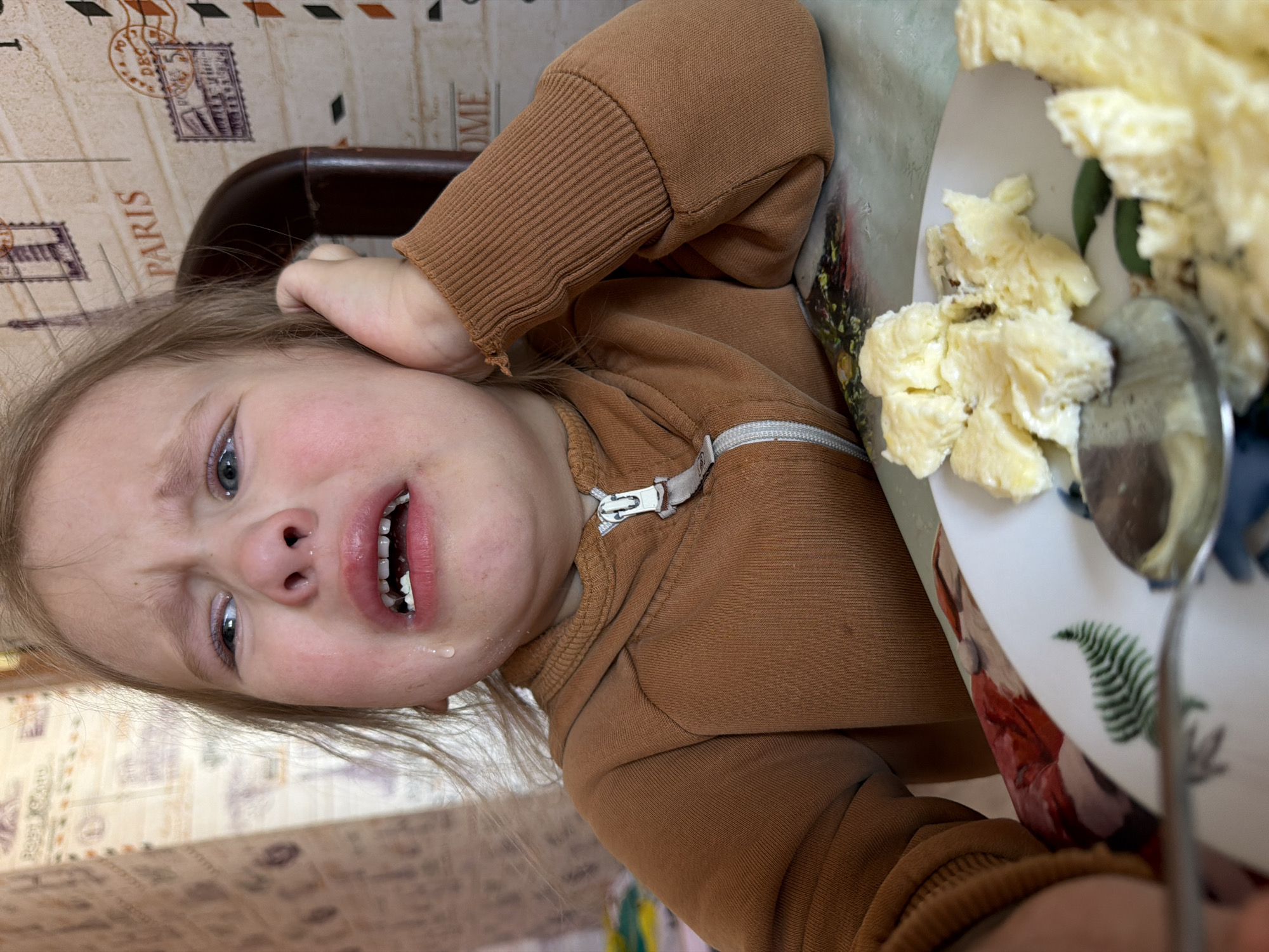 The image shows a child with fair skin and light hair in distress, likely crying, with tears visible on their face. They are wearing a brown zip-up jacket. A plate of scrambled eggs is nearby, along with a spoon. The background features a patterned table surface and a chair back. The location is Chaikovsky, Russia.