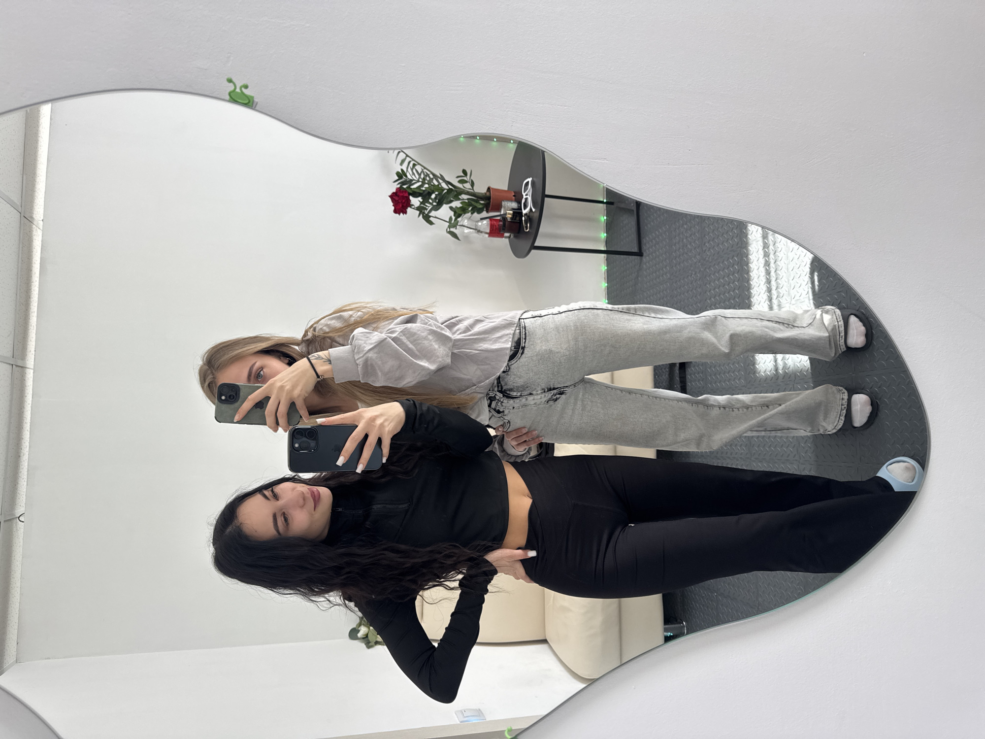 Two young women are reflected in an oddly shaped mirror, taking a selfie with their phones. The woman in the foreground is wearing a black long-sleeved top and black pants, while the other woman is wearing a light gray sweatshirt and gray jeans. The background features a white wall, a gray-tiled floor, a small black table with a potted plant and other items, and a beige sofa. There is a green clip-like object attached to the top of the mirror frame. I cannot confirm the location as Фоки, Russia, based on the image alone.