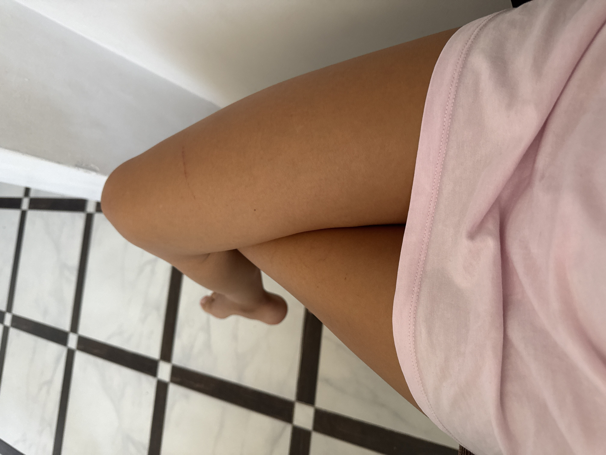 An over-the-shoulder shot captures a person's legs, one crossed over the other, in an indoor setting. The individual is wearing a light pink t-shirt. The floor features a geometric pattern of alternating black and white squares, possibly marble tiles. To the right, a white wall creates a backdrop. A faint reddish mark is visible on the upper portion of the visible leg. The lighting suggests it is daytime. There are no identifiable landmarks or specific elements that can definitively place the scene within Chișinău, Moldova.