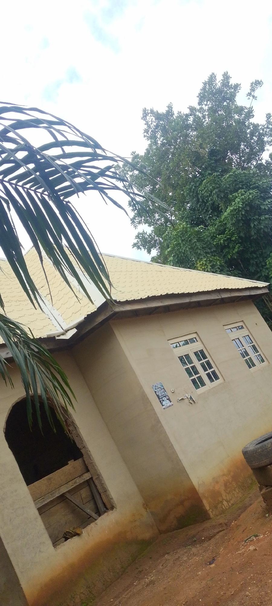 This media file displays an outdoor scene in Baissa, Nigeria. The primary subject is a single-story house with a light yellow, corrugated metal roof. The house has a beige, stucco-like exterior. Two windows with white frames and multi-pane glass are visible on the side of the house. To the left, there is an arched opening that appears to be an unfinished or unglazed window, boarded up with wooden planks. A small poster or flyer is affixed to the wall between the opening and the windows.
In the foreground, a large palm frond extends into the frame from the left, casting shadows on the roof. Behind the house, a dense canopy of green trees fills the background. The sky is overcast with visible clouds, suggesting it might be daytime, possibly morning or afternoon, with diffused light. The ground in front of the house is a reddish-brown dirt path. A discarded tire is stacked on top of another object near the right edge of the frame. The overall impression is that of a rural or semi-rural residential setting. There is no discernible activity taking place; the image appears to be a still photograph of the house and its surroundings.