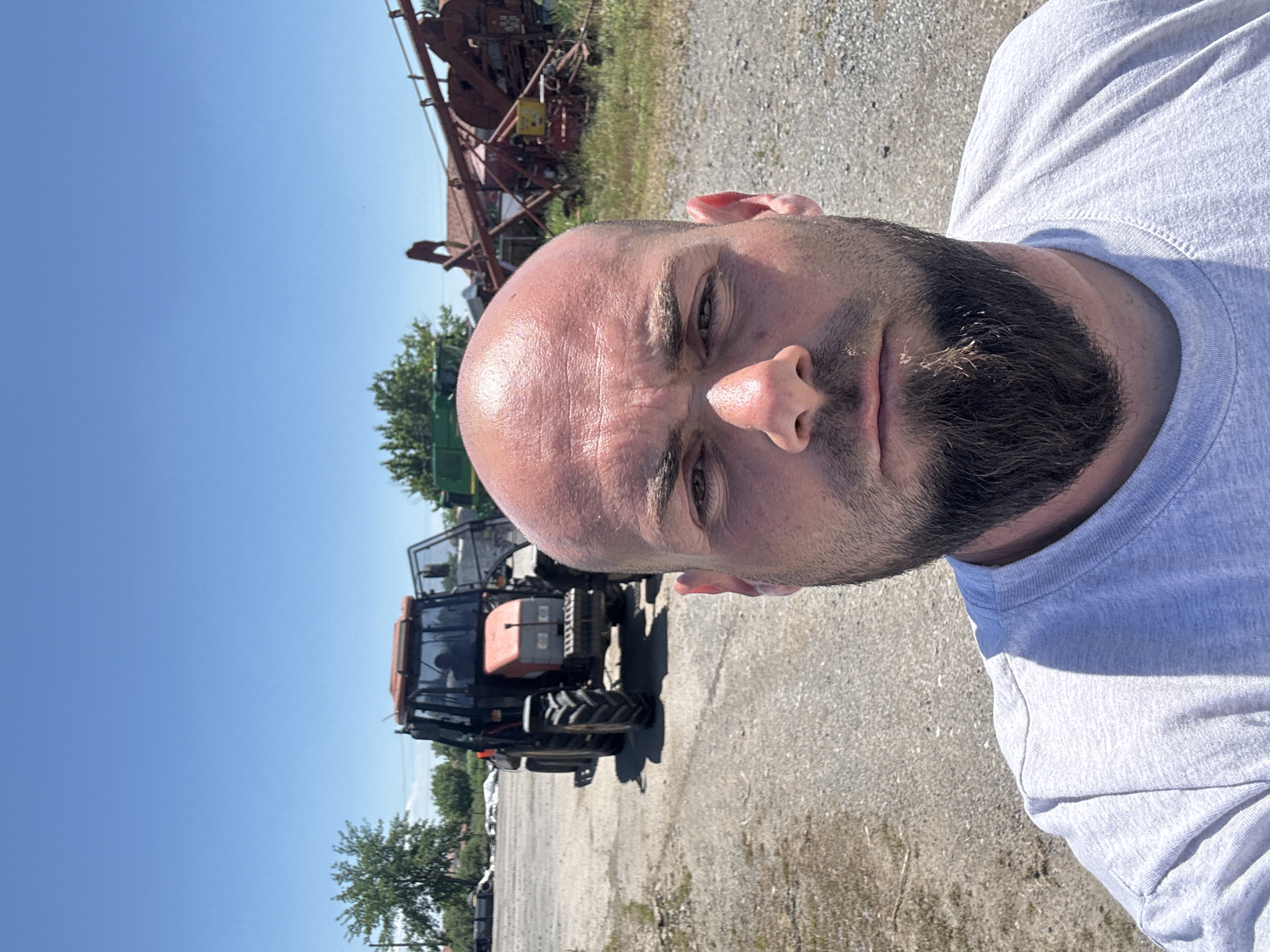 A close-up photo of a bald man with a beard, wearing a gray t-shirt, likely outdoors. The background includes agricultural machinery and a gravel ground cover, typical of a rural setting, the scene appears to be shot in an outdoor environment, given the bright natural light. No specific details confirm the location as Sarnevo, Bulgaria, but the agricultural equipment suggests a rural area.