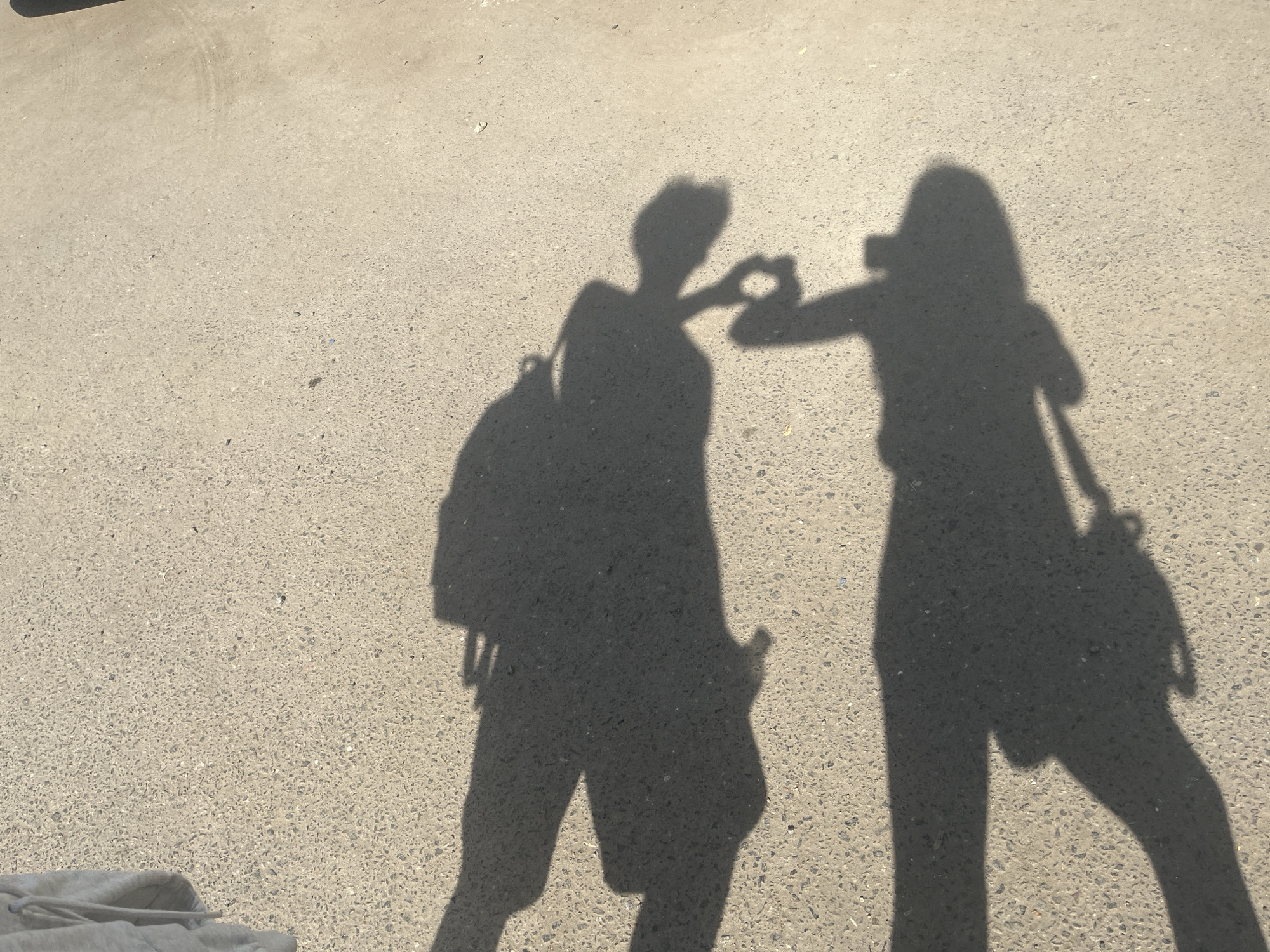 The image displays the shadows of two individuals interacting on a light-colored, granular paved surface, likely asphalt or concrete, under bright sunlight. The scene is outdoors, consistent with a street, sidewalk, or open plaza in Unknown, Unknown.
On the left, the shadow of a person is visible, wearing a backpack and appearing to have relatively shorter hair. Their right arm is extended forward, with the hand curved to form half of a heart shape. On the right, the shadow of a second person is visible, carrying a bag with a visible strap over their left shoulder/arm and appearing to have longer hair. Their left arm is extended forward, with the hand curved to complete the heart shape formed with the first person's hand. Both shadows are facing each other in the upper-middle section of the frame. The paved surface is speckled with small dark aggregates. A light-colored fabric item is partially visible in the bottom-left corner of the frame. The strong, distinct shadows indicate a high sun angle.
