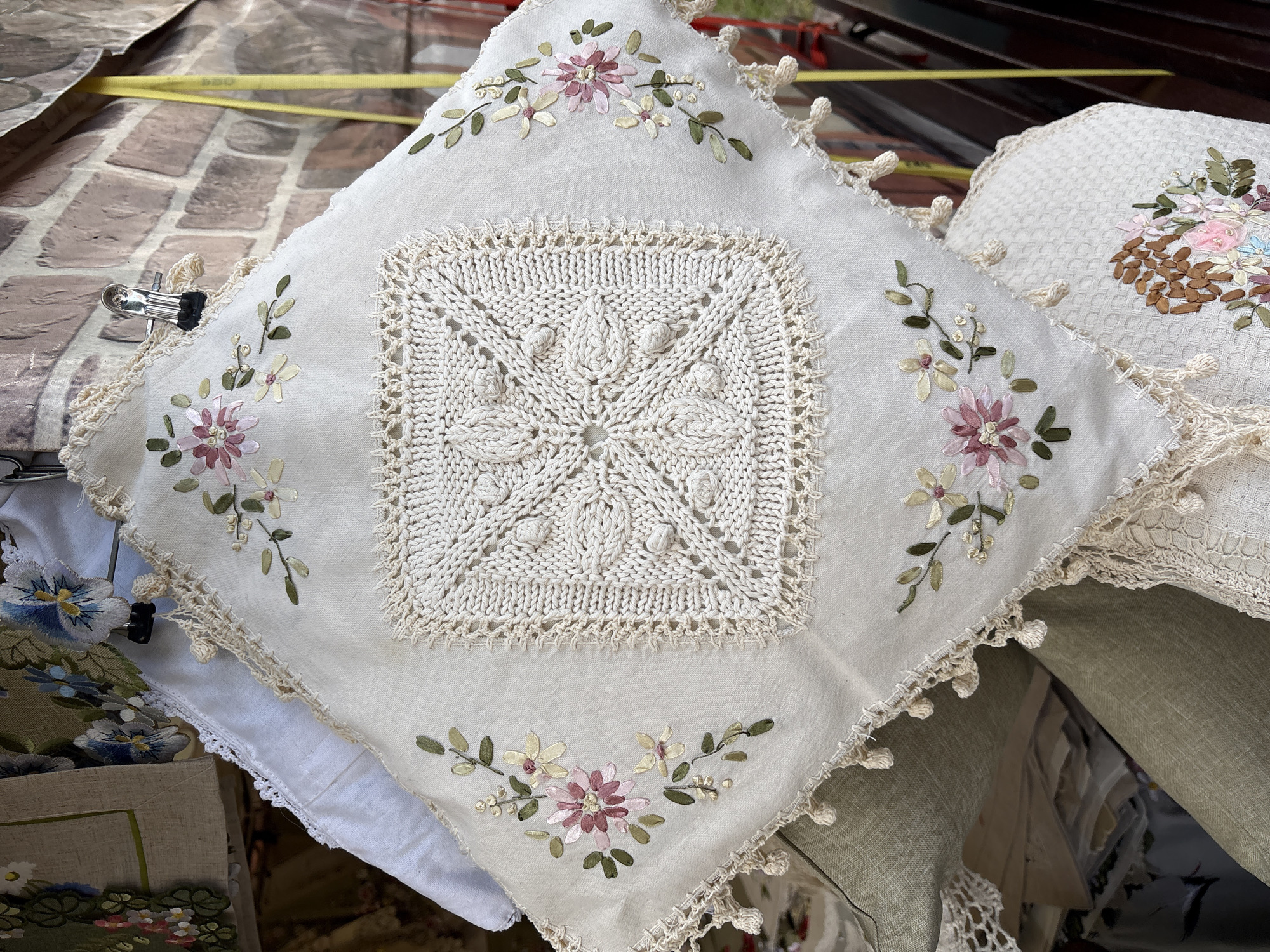 The image shows a close-up of a decorative pillow with intricate embroidery and crochet work. The pillow is light-colored, possibly linen or cotton, and features floral motifs embroidered with ribbon in shades of pink, white, and green. The center of the pillow has a square crocheted panel with a starburst pattern. The edges of the pillow are adorned with a delicate crocheted trim and small tassels. The pillow appears to be displayed outdoors, possibly at a market or craft fair, given the brick wall in the background and the hint of yellow tape visible. The overall aesthetic suggests a handcrafted, artisanal item.
