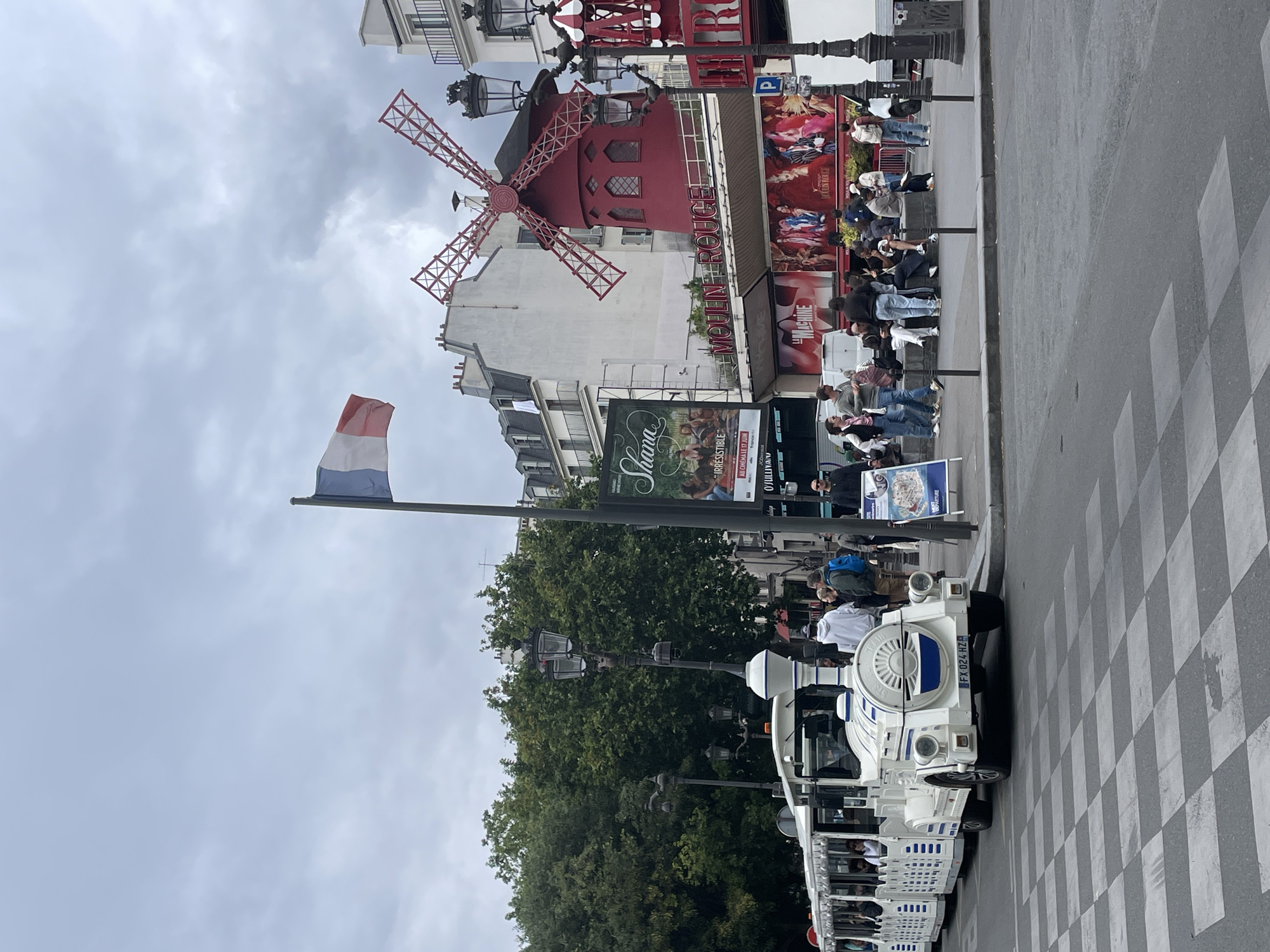 This is a medium shot photograph taken outdoors, likely during the daytime, with overcast skies suggesting a cloudy or perhaps drizzly day. The scene is set in Paris, France, featuring the iconic Moulin Rouge windmill as a prominent landmark.
In the foreground, a white tourist train with open-air seating is parked on a street next to a pedestrian crossing. Several people are visible inside the train, suggesting a tour or excursion is underway. A French flag flutters on a flagpole to the left of the Moulin Rouge.
The Moulin Rouge building itself is painted red and is adorned with its distinctive red windmill. Posters advertising shows are displayed on the exterior of the building. A crowd of people is gathered in front of the Moulin Rouge, possibly waiting to enter or observe the landmark. The overall atmosphere appears to be one of tourism and sightseeing.
Visible text includes "MOULIN ROUGE," "FRANCE," "MACHINE," "IRRESISTIBLE," and a license plate reading "FX 024-HZ." The cloudy weather contributes to a subdued lighting condition, without strong shadows. The focus is on the interplay between the tourist activity, the historic landmark, and the urban environment.