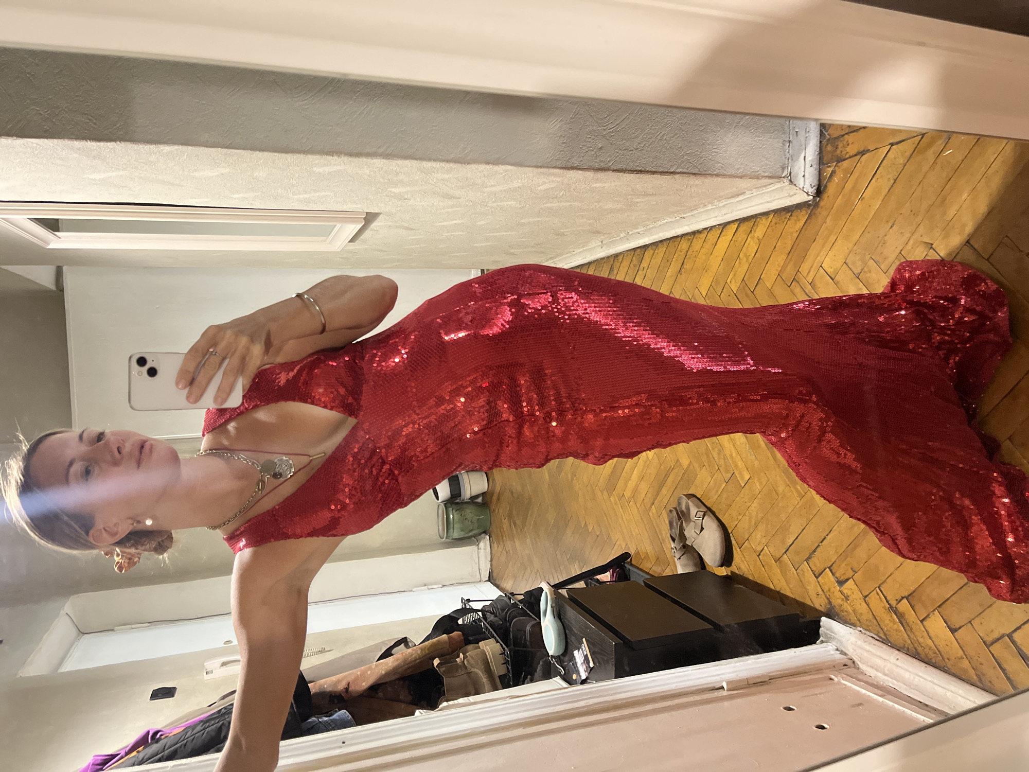 In a well-lit indoor setting, a person is reflected in a mirror, wearing a striking, form-fitting, floor-length red sequined dress. The dress features long sleeves and a deep V-neckline, accentuating the wearer's silhouette. A silver necklace with a pendant is visible around their neck. The person is holding a smartphone, presumably to take the photograph, and is looking towards the mirror. Their left arm is bent, with their hand resting near their head, and they are wearing a thin bracelet on their wrist. The background of the reflection shows a room with wooden parquet flooring and some furniture, including a black cabinet and a rack with various items. A pair of light-colored shoes are visible near the cabinet. The overall impression is of someone posing for a photo in a dressy outfit.