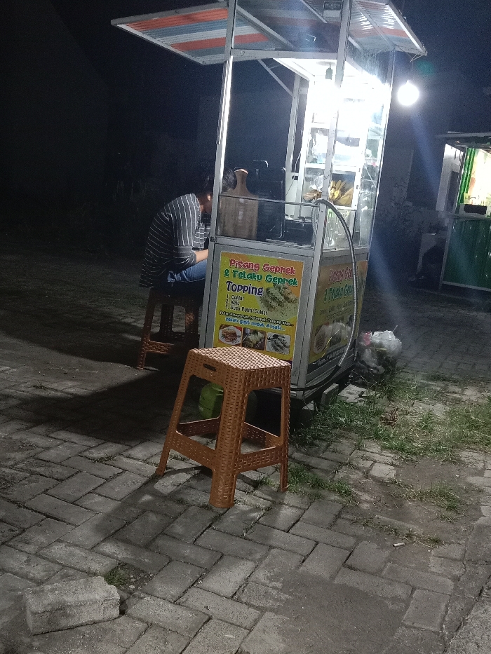 This image captures a nighttime scene in the city of Turunsih, Indonesia, depicting a street food vendor setup. A brightly illuminated food cart, constructed from silver-colored metal and clear panels, stands on a grey herringbone-patterned brick pavement. The cart features a red, white, and blue striped awning and displays a prominent yellow sign advertising "Pisang Geprek & Telaku Geprek TOPPING" with various options listed and food images. Internal lights illuminate the cart's contents, and an external bare bulb hangs from its frame, casting shadows.

One individual, wearing a dark and light striped long-sleeve shirt and dark trousers, is seated on a brown woven plastic stool behind the cart, facing away from the camera and leaning forward. An identical empty brown woven plastic stool is positioned directly in front of the cart. To the right of the cart, the brick pavement transitions to a patch of sparse grass, where a white plastic bag, presumably containing refuse, and other small debris are visible. A single grey concrete block rests on the paved ground in the lower-left corner. In the background, a less illuminated, green-paneled stall or structure is partially visible to the far right, set against a dark, indistinct backdrop indicating an outdoor, unlit environment beyond the immediate vendor area.