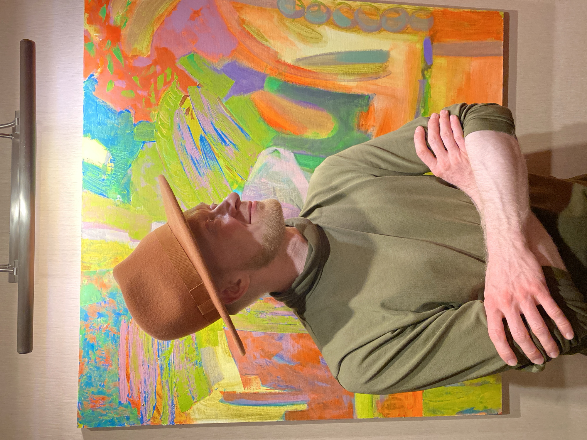 The image depicts a man with a beard, wearing a brown hat and an olive green long-sleeved shirt and pants. He is standing in front of a colorful abstract painting that has shades of green, blue, orange, and purple. His arms are crossed and he is looking upwards. To the left is a vertical light fixture. The setting appears to be indoors. The city of Yekaterinburg, Russia, is not discernible from the image alone.