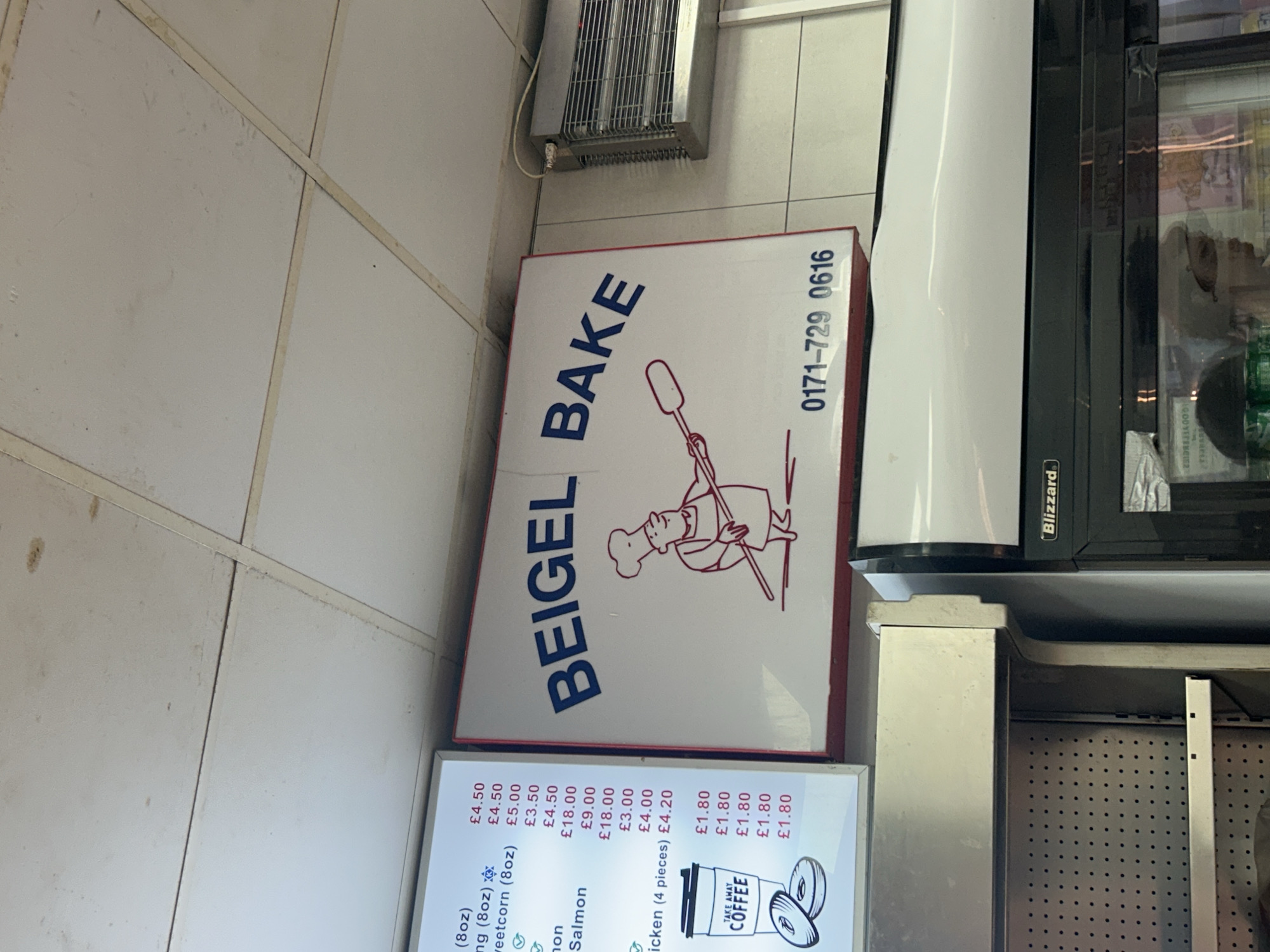The image shows a close-up, eye-level view of a "BEIGEL BAKE" shop sign, which is white with red trim and blue lettering. A cartoon chef is depicted on the sign, holding a paddle. Below the cartoon chef, a phone number "0171-729 0616" is displayed. To the left of the "BEIGEL BAKE" sign, a menu board lists various items and prices. The menu includes items like "corn (8oz)" priced at £4.50, "sweetcorn (8oz)" at £5.00, and "Chicken (4 pieces)" at £4.20. There is also an illustration of a coffee cup and two bagels, with "TAKE AWAY COFFEE" written on the cup. Prices for coffee are listed as £1.80. The shop appears to be located in London, United Kingdom, based on the pricing currency (£) and the commonality of "Beigel Bake" establishments in the city. The background shows a white tiled wall and a fluorescent light fixture on the ceiling. To the right of the sign is a silver heating element attached to the wall. Below the sign, a portion of a white and black refrigerated display case labeled "Blizzard" is visible. A person wearing a hairnet or cap is reflected in the glass of the display case. The overall environment suggests an indoor food establishment.