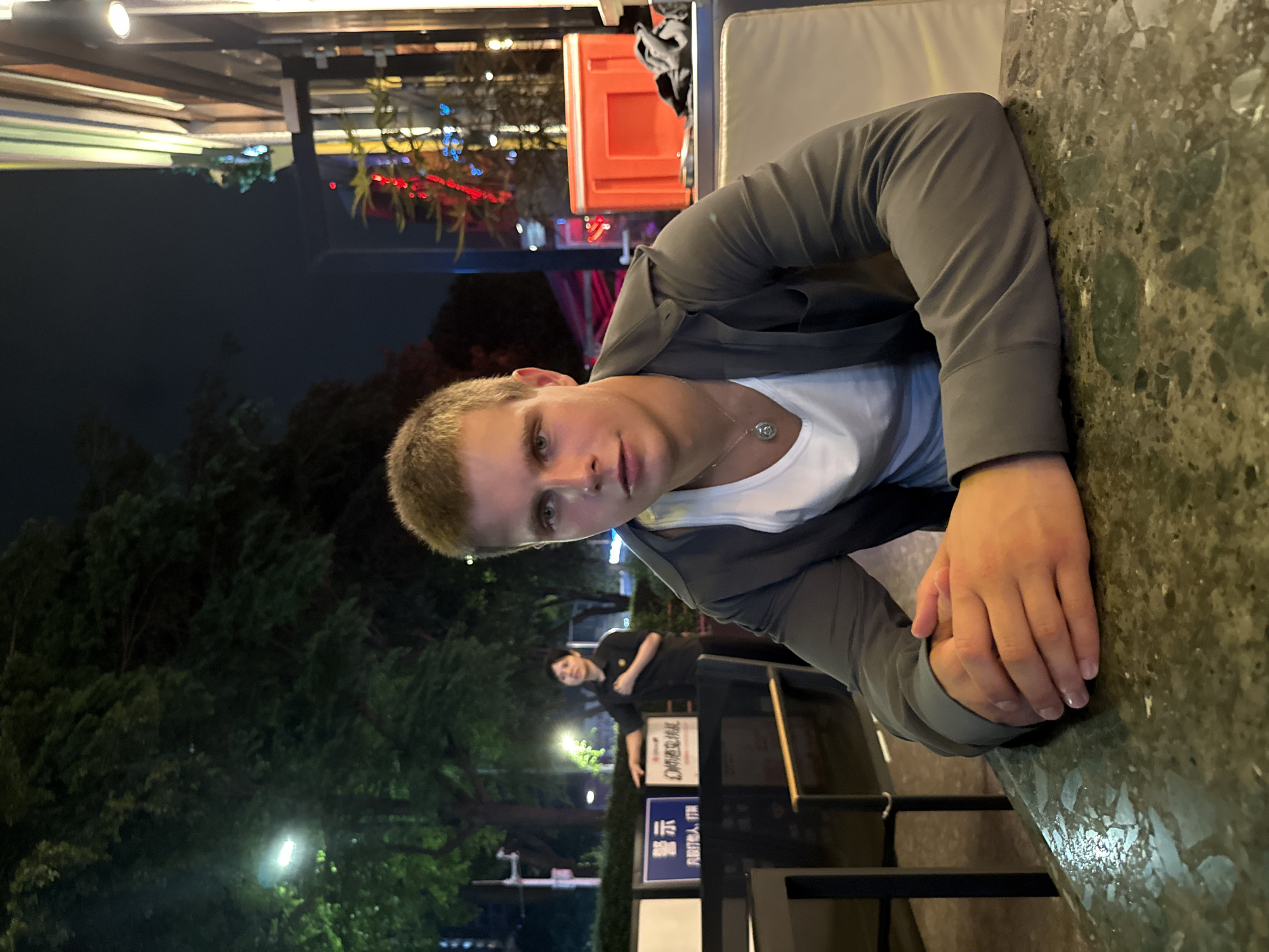 A person with short blonde hair wearing a gray jacket and white t-shirt sits at a table, resting their hands on the surface. The location appears to be outdoors at night in Tianhe District, China, based on the signage visible in the background. Trees and streetlights can be seen behind the person. Another individual is visible standing further back, to the person's left.