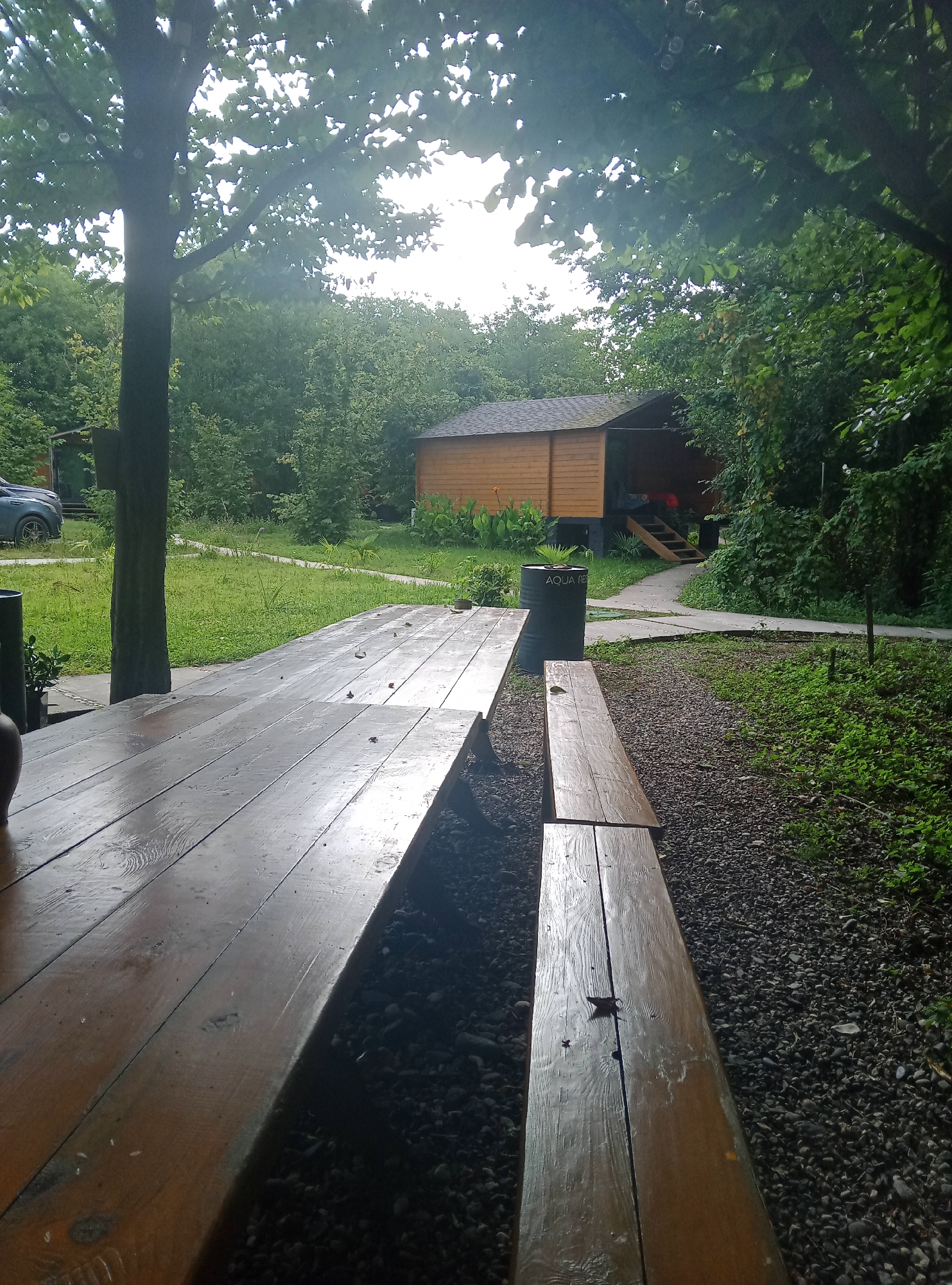 In Pshap, Abkhazia, a wooden picnic table and bench sit outdoors on a graveled area, partially shaded by trees. A wooden structure, possibly a cabin or shed, is visible in the background, with steps leading up to its entrance. To the left of the picnic table, a dark-colored vehicle is parked on a grassy area. The scene appears to be a recreational or resting spot, with lush greenery surrounding the area.